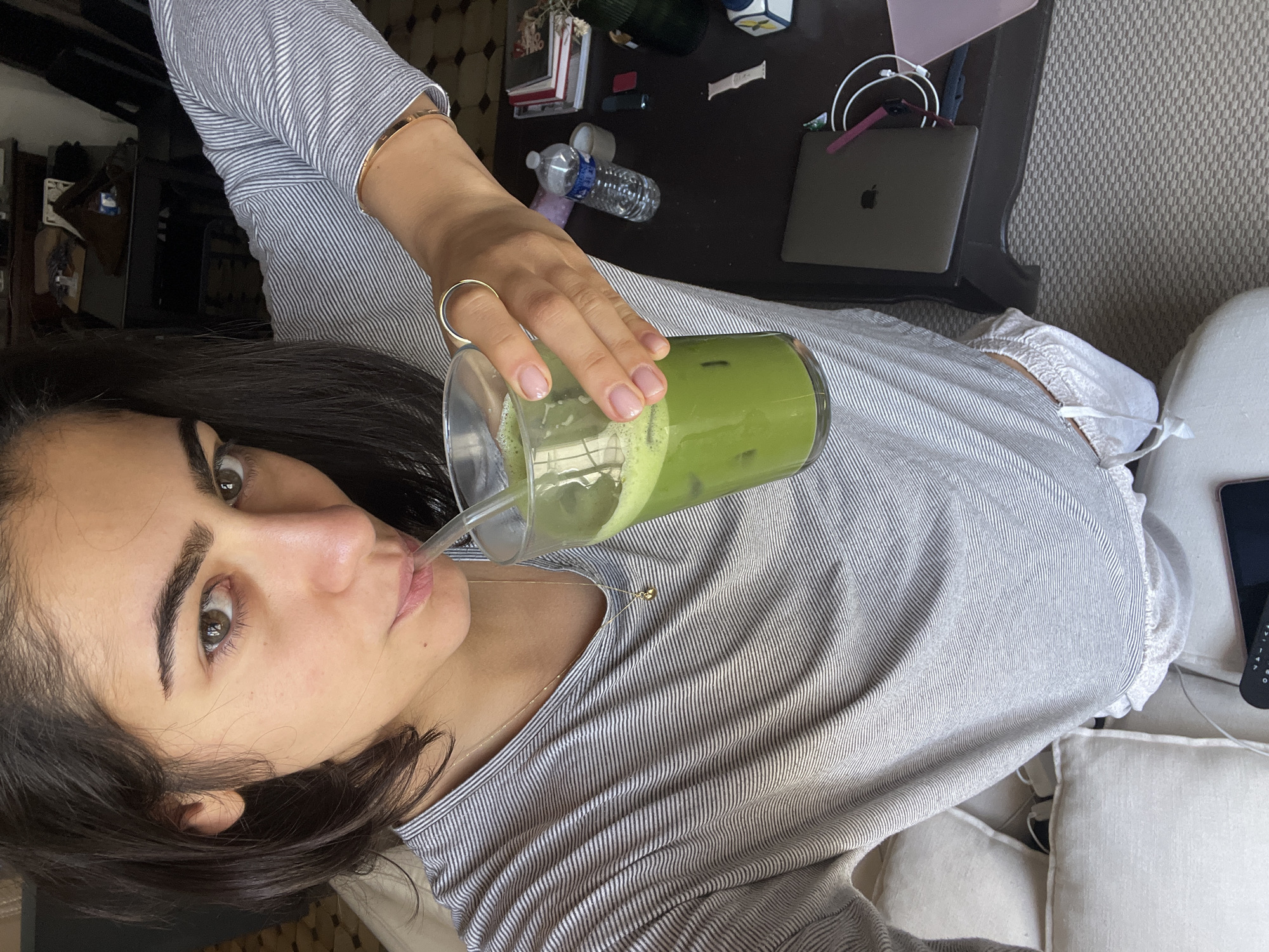 A woman is drinking a green beverage with ice through a clear straw. She is sitting on a white couch, wearing a striped shirt and has a gold ring on her finger. A black desk is nearby holding a laptop, a water bottle, and other items. The location is likely in Paris, France, as indicated by the architectural details and ambiance of the scene.