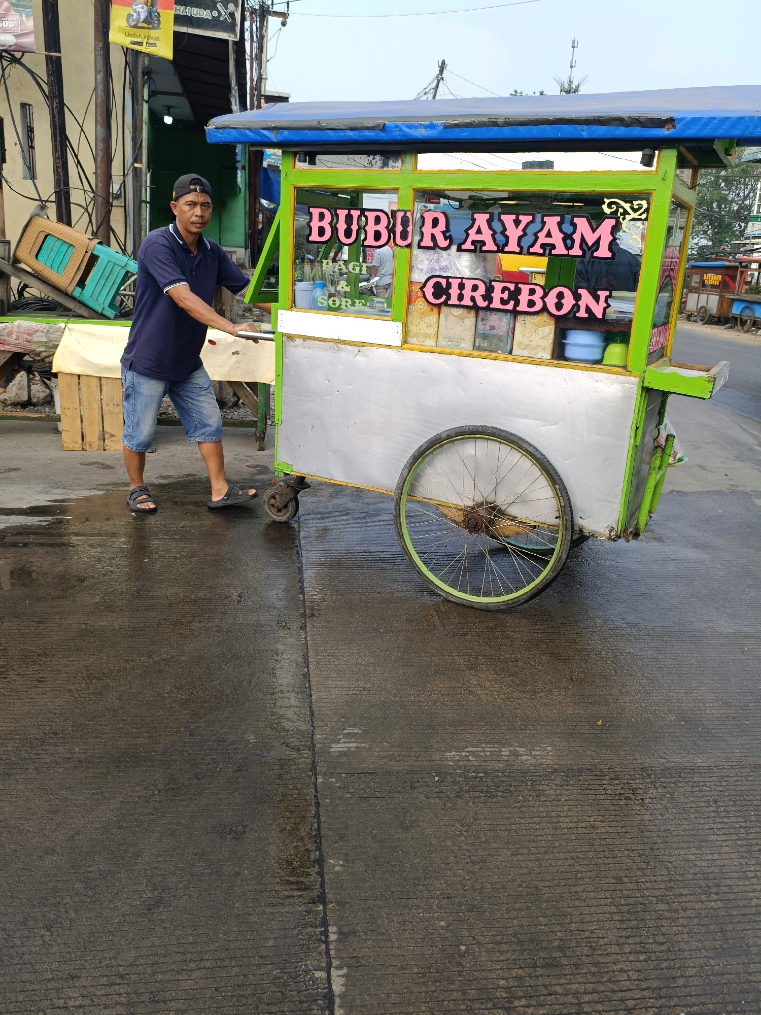 A man stands beside a food cart selling "Bubur Ayam Cirebon" in Tangerang, Indonesia. The cart is green and silver, with the name of the food in pink lettering on the front. The man is wearing a dark blue polo shirt and denim shorts. He is holding onto the cart, as if about to push it. The cart has a large wheel on the side. The ground is wet, suggesting it may have recently rained. In the background, there are buildings and other carts.