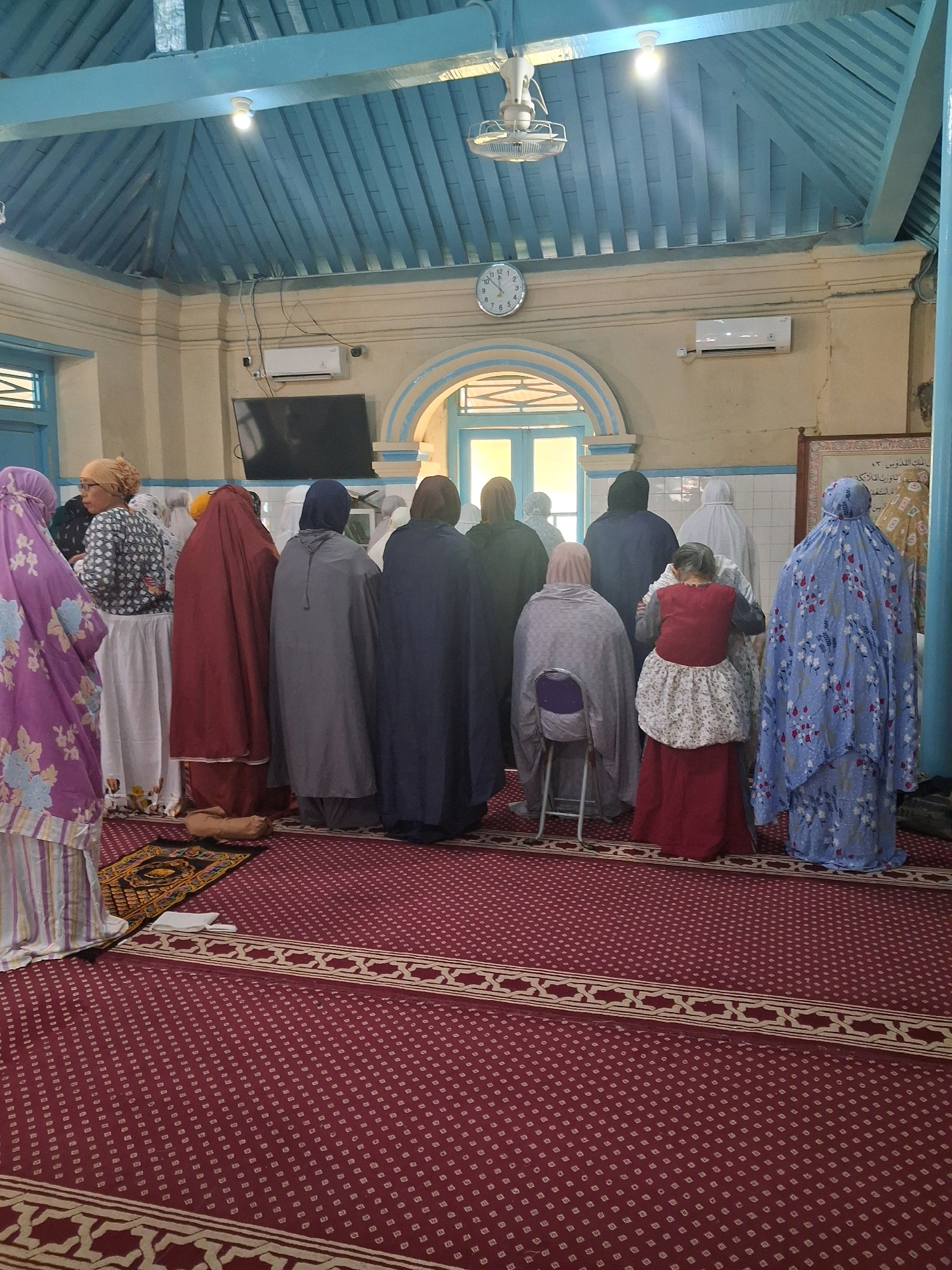 In the city of Surakarta, Indonesia, a group of women are gathered in prayer inside a room with a blue, sloped ceiling supported by wooden beams. The walls are a pale yellow, and an arched doorway with light blue trim is visible in the background. A black television is mounted on the wall to the left, and an air conditioning unit is above it. A round clock with Roman numerals hangs on the wall in the center of the room. The floor is covered with a red carpet featuring a repeating pattern of small squares and a decorative border. The women, all dressed in various religious garments covering their heads and bodies, are facing away from the camera, some standing and some kneeling or seated on a chair. One woman in the foreground is wearing a vibrant purple and pink patterned dress. To her left, another woman wears a patterned gray and white top with a white skirt. Further into the scene, women are seen in burgundy, gray, dark blue, and light blue robes. One woman in a red dress with a white patterned apron is seated on a metal chair. A prayer mat is placed on the carpet in the foreground. The overall scene depicts a moment of communal prayer within a religious or community space in Surakarta.
