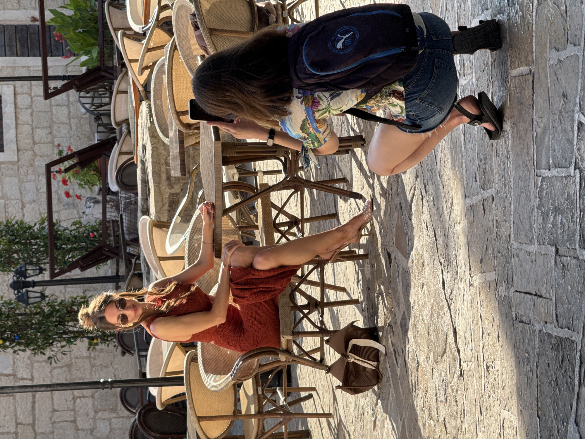 An outdoor cafe scene in Budva, Montenegro, features a woman in a red dress posing while seated at a table. She wears sunglasses and her hair is styled in a bun. A brown handbag rests on the ground beside her chair. In the foreground, a second woman kneels, holding a smartphone to take a photograph of the seated woman. This second woman wears a floral print shirt, denim shorts, and a backpack. The background consists of numerous empty outdoor cafe chairs and tables, with a stone building wall visible behind them. The ground is paved with flagstones.