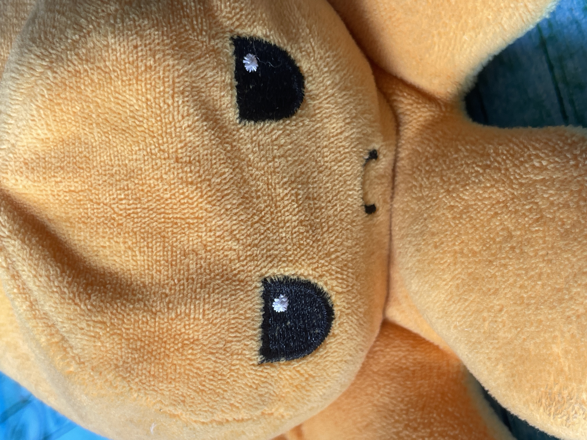 The image is a close-up shot of an orange plush toy with black eyes and a small mouth. The toy appears to be a character, possibly an octopus due to the shape of the head and visible tentacles. The texture of the plush material is clearly visible. The background is out of focus but appears to be a blue surface with a wood grain pattern. There are no people or specific locations in Tashkent, Uzbekistan identifiable in this image.