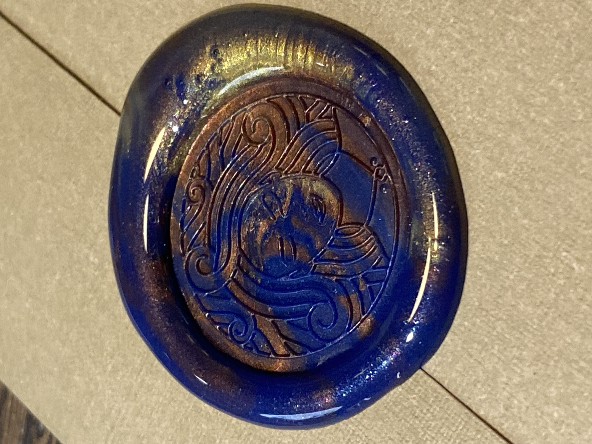 A close-up shot shows a circular wax seal, predominantly blue with gold and brown highlights. The seal features an embossed design, seemingly of a woman's face and flowing hair, encircled by an ornate border. The wax appears glossy and slightly irregular in shape. The seal rests on a light brown, textured surface. There is no indication that this image is located in the city of Votkinsk, Russia.