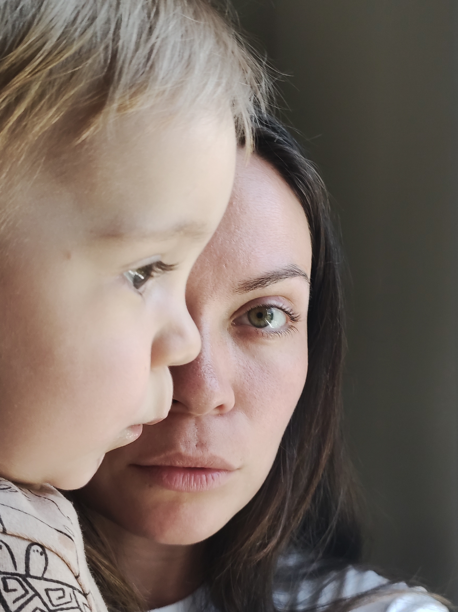 A close-up shot shows a fair-skinned woman with dark brown hair partially framing her face, situated beside a fair-skinned child with light blonde hair. The woman's face is predominantly visible; she gazes forward with an intent expression, her green eye is clear and focused. The child is positioned to the left of the woman, with only the side of their face visible; they appear to be looking away from the viewer. The background is out of focus, with a dark, undefined area behind them. The child is wearing a light-colored garment with dark, geometric patterns. The setting appears to be indoors, near a window, though the specific city and location are not determinable from the image.
