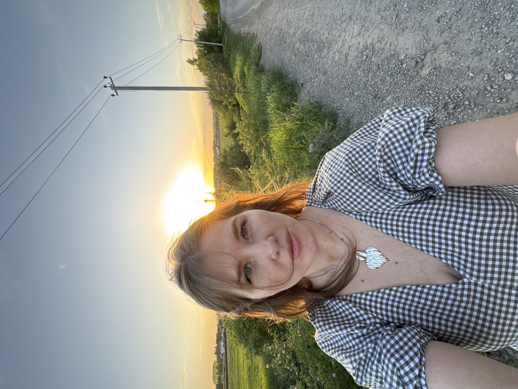 A medium-shot photograph captures a young woman outdoors against a vibrant sunset backdrop. She is wearing a green and white checkered dress paired with a silver necklace featuring a leaf-shaped pendant. The setting sun casts a warm glow on her face, with the sky transitioning from a clear blue to orange. In the background, an overhead power line stretches across the frame, indicating the presence of human infrastructure. The foreground consists of a gravel path and grassy vegetation, suggestive of a rural or semi-rural location in Горный, Russia.