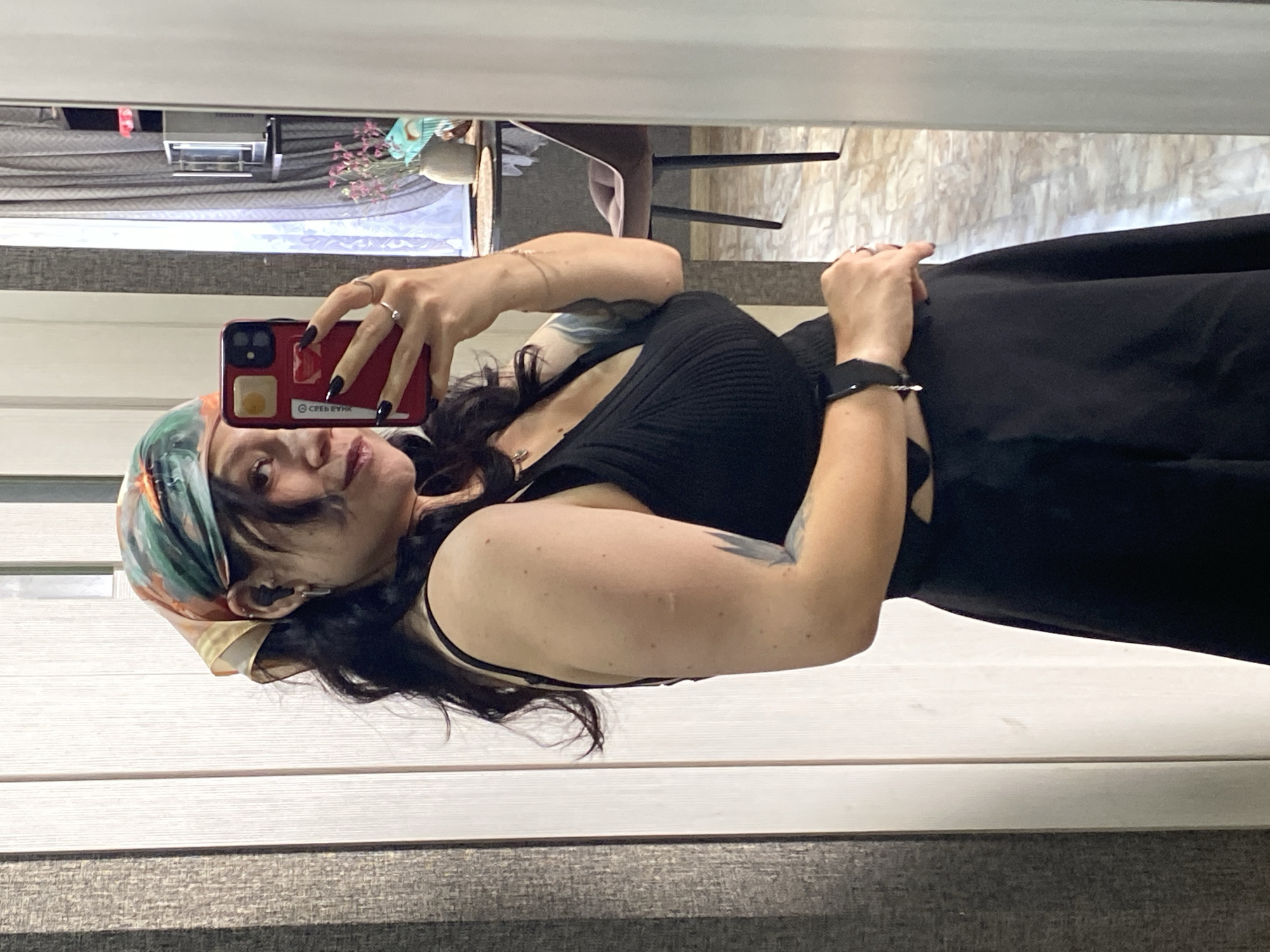 A woman is taking a selfie in a mirror. She is wearing a black dress and a colorful headscarf. Her dark hair is pulled back and falls over her shoulders. She has dark nail polish and is wearing rings on her fingers. The background appears to be a room with vertical blinds and a mirror. In the reflection of the mirror, there is a dining table and chairs, as well as a vase with flowers. There are also some items on shelves. The overall impression is that the woman is in a residential setting.