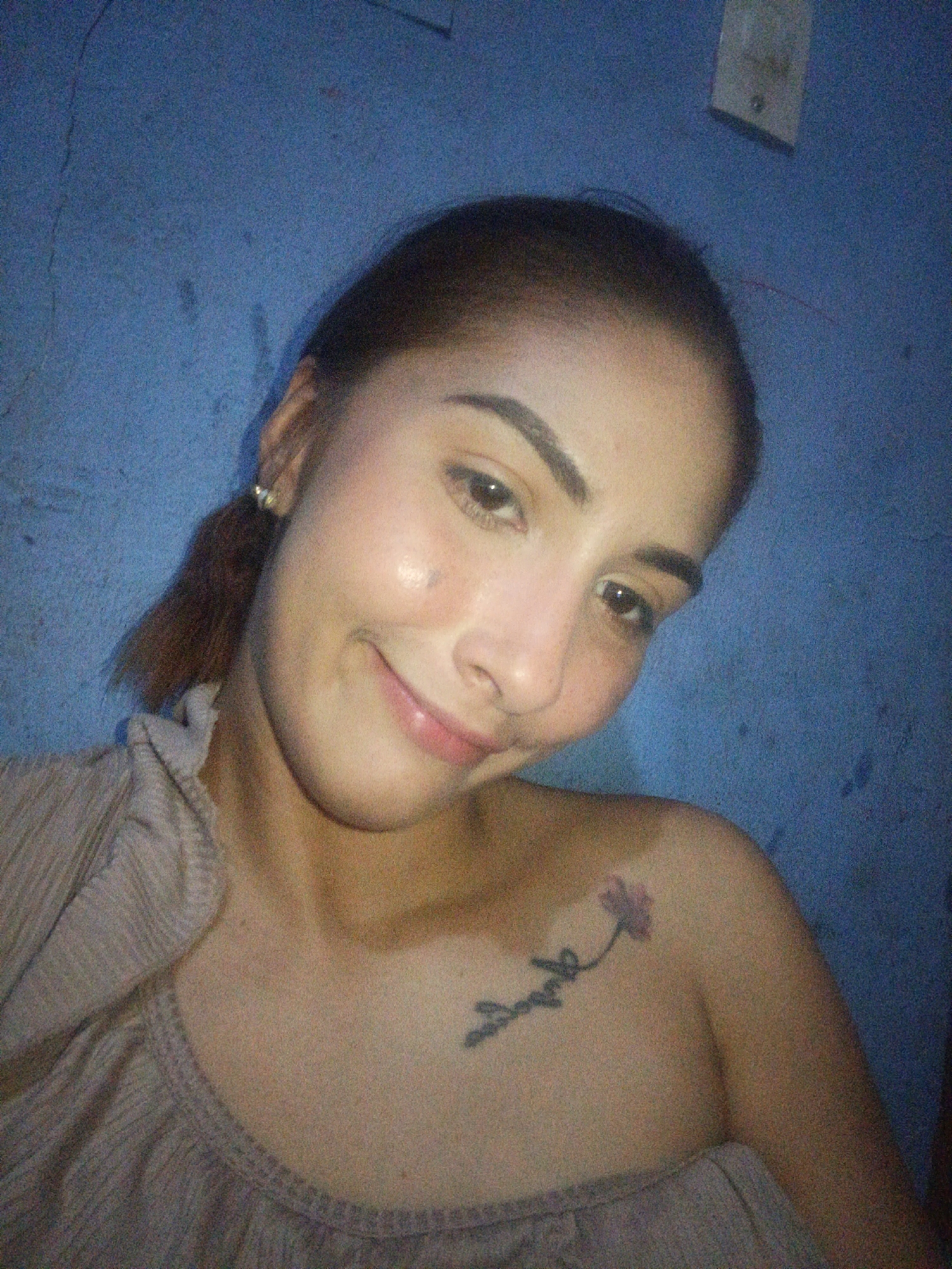 A close-up photograph depicts a woman with light skin, brown hair pulled back in a ponytail, and a slight smile. She is wearing a light-colored, ruffled top. A floral tattoo is visible on her upper left chest. The background is a blue wall. A white electrical fixture is visible in the upper right corner of the frame. There is no information to indicate the location in the city of Juan Griego, Venezuela, or any actions, interactions, or any other subjects and objects.