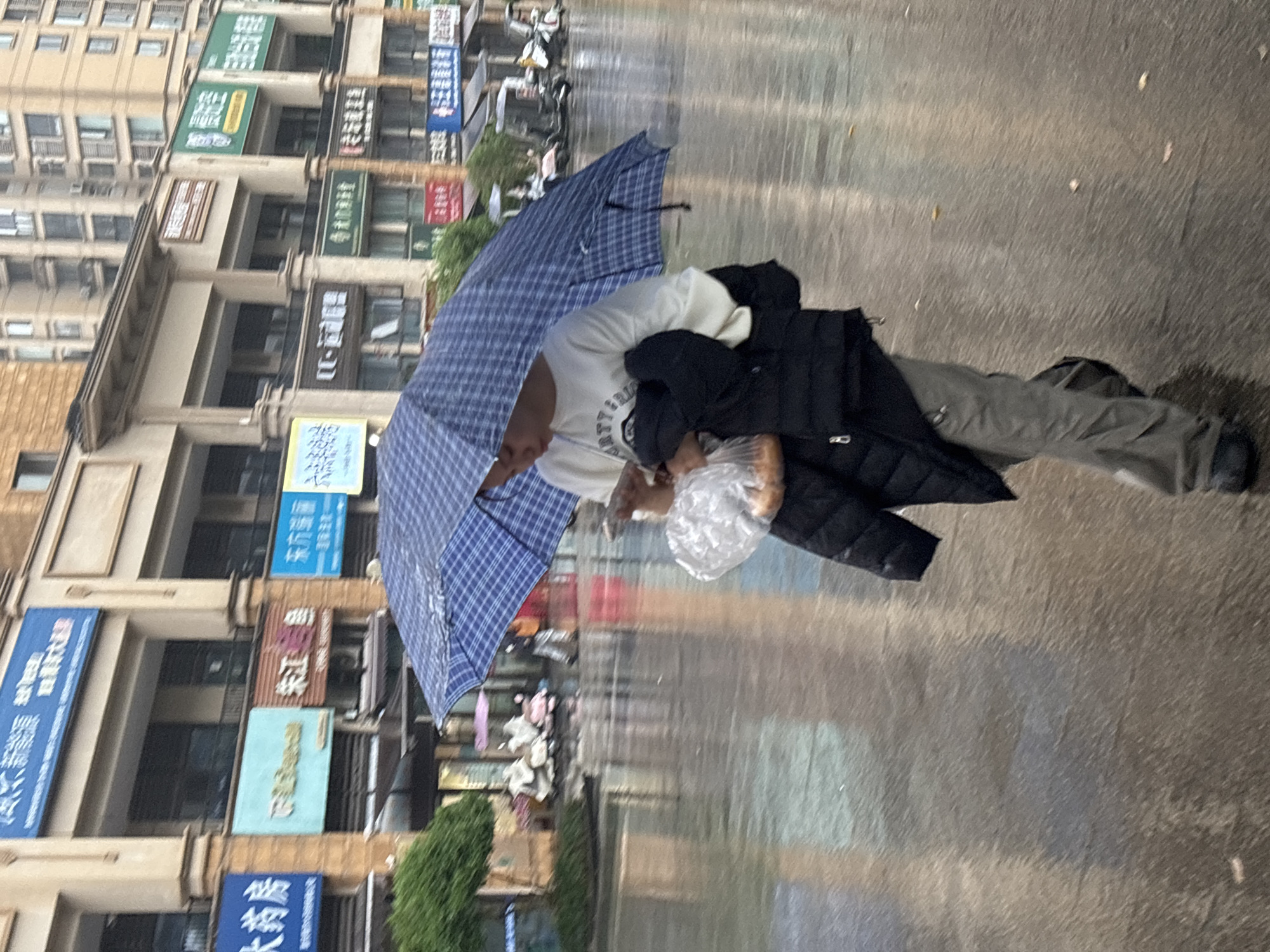 In Jiangjin, China, a person with dark hair walks on a wet city street holding a blue and white checkered umbrella. The individual wears a light-colored sweatshirt, dark pants, and a black jacket. They hold a clear plastic bag containing bread. Behind them are buildings with signs written in Chinese characters. The street is slick with rain. There are parked scooters.