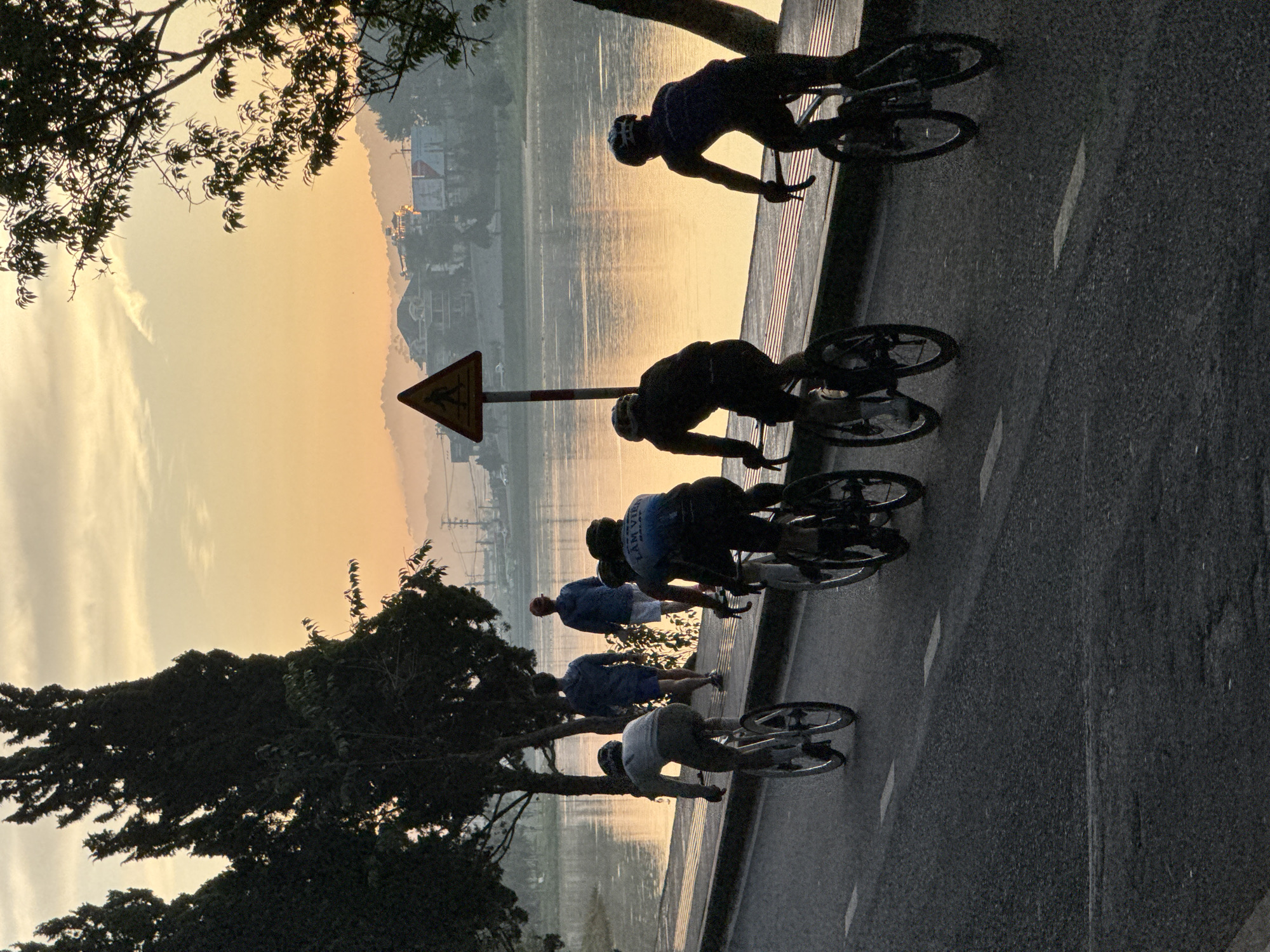 A group of five cyclists are riding on a paved road alongside a body of water, likely Xuan Huong Lake, in Da Lat, Vietnam, during sunrise. The sky is a warm gradient of yellow and orange, with some wispy clouds. The cyclists, silhouetted against the bright sky, are positioned from left to right across the frame. The first cyclist on the far left is wearing a light-colored top and dark shorts. Next are two cyclists in dark cycling attire. The fourth cyclist is in the foreground, also in dark attire. The fifth cyclist on the far right is wearing a dark top and helmet. Two individuals are walking on the paved walkway between the road and the water. A triangular pedestrian crossing sign is visible on the right side of the walkway. Tall, dark green trees line the left side of the road. The water is calm, reflecting the colors of the sky. In the background, rolling hills with scattered buildings are visible through a light mist. The road has visible white lane markings.