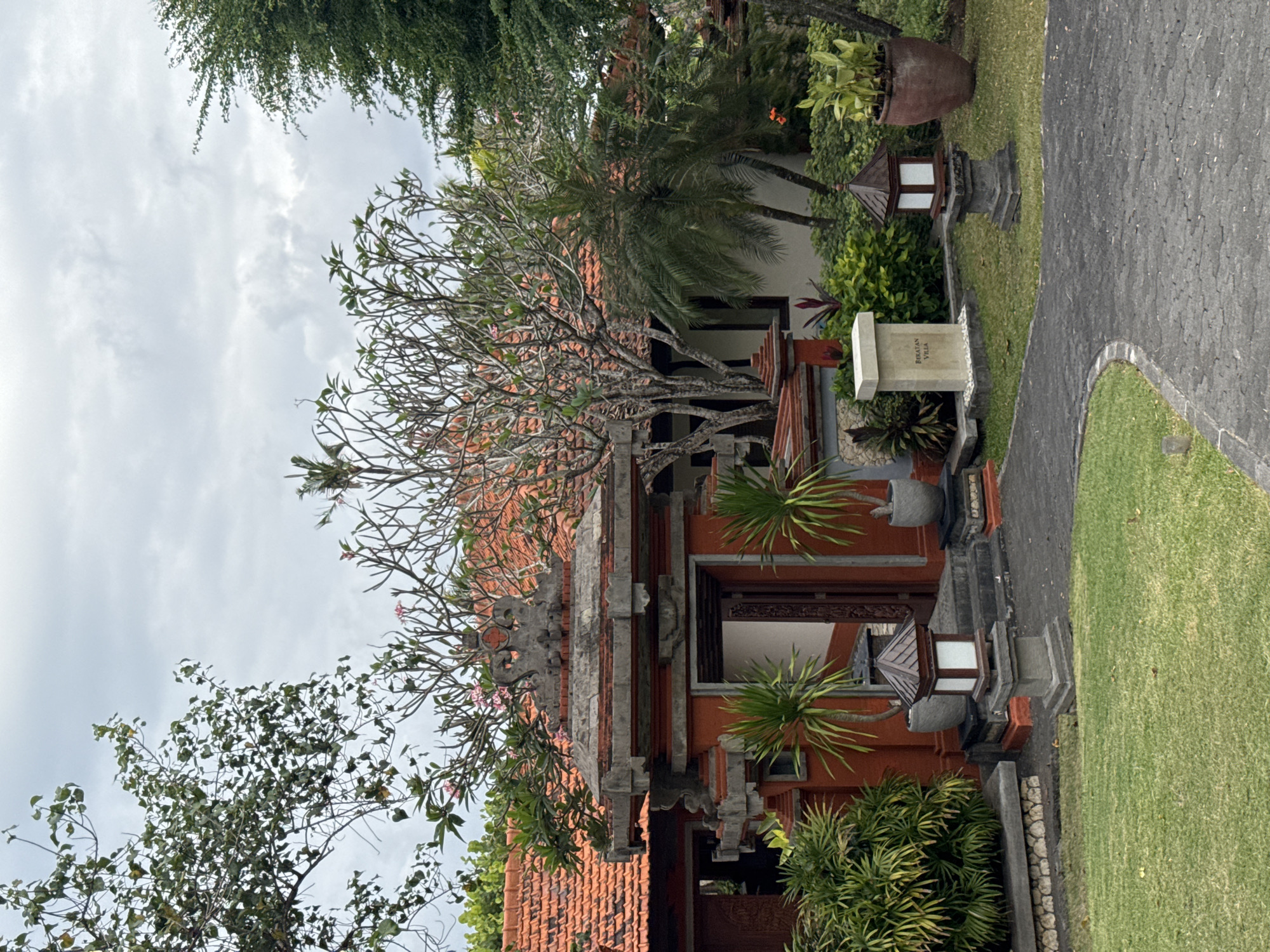 An outdoor medium shot captures a pathway leading towards a traditional Balinese entranceway. The entrance is adorned with intricate stone carvings and a vibrant orange-red paint. Flanking the pathway are meticulously maintained green grass lawns and various lush plants, including palm trees and potted flora. The background reveals a building with an orange tiled roof, partially obscured by leafy trees under an overcast sky. Two stone lanterns and a memorial marker with the inscription "BERATAN VILLA" are visible along the pathway, suggesting a residential or resort setting in Kuta Selatan, Indonesia. The scene depicts a tranquil and culturally rich environment.