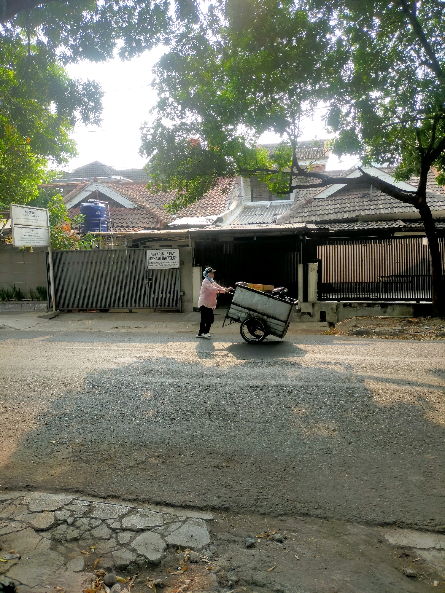 A person is pushing a cart down a street lined with houses and trees in Bandung City, Indonesia. The person, wearing a pink shirt, black pants, and a light-colored head covering, is facing away from the camera. The cart appears to be a wheeled utility cart. The houses have tiled roofs and some have fences. The street is paved with asphalt and there are shadows from the trees on the ground. The sky is bright and appears to be daytime.