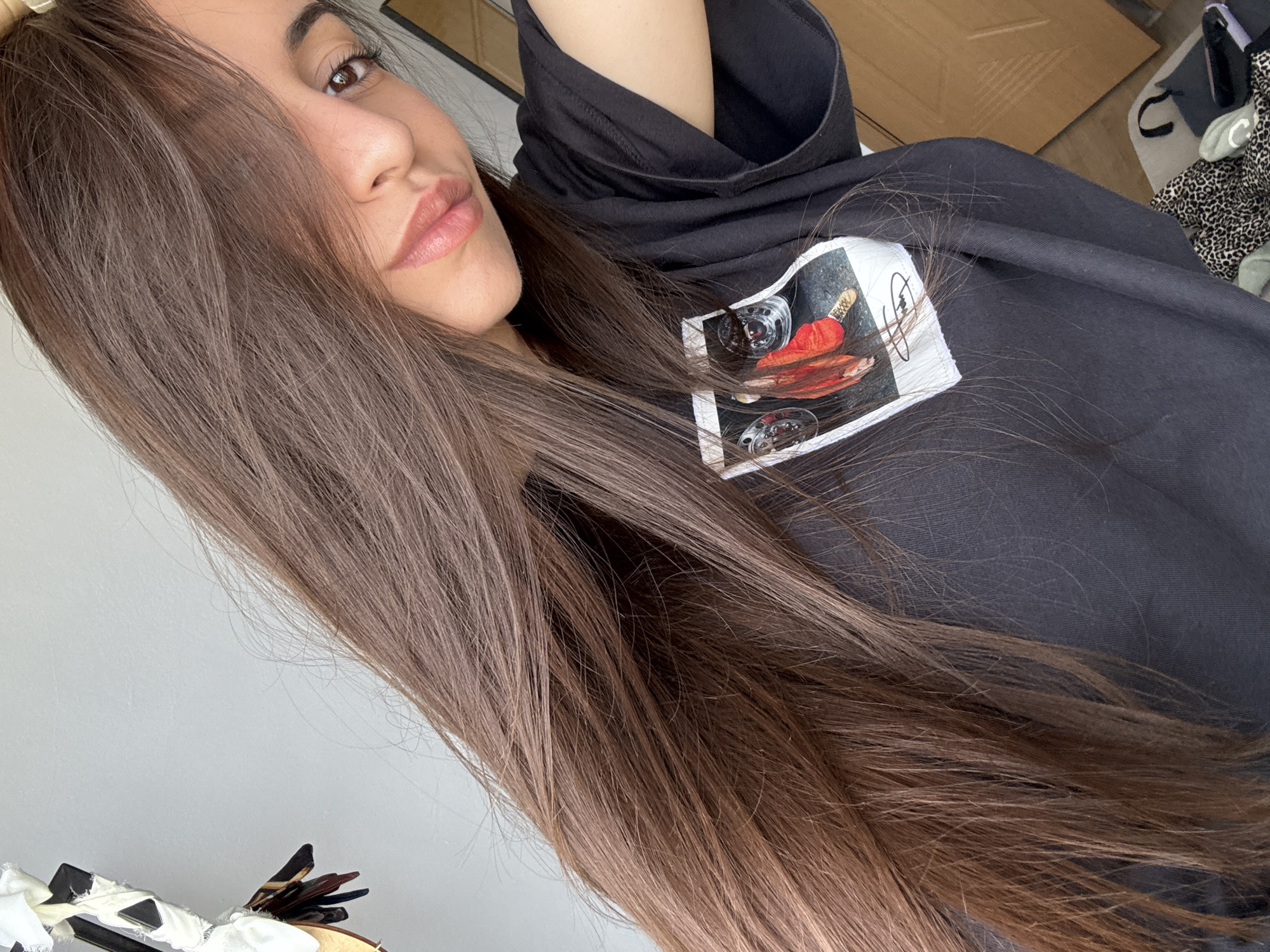 A medium shot shows a woman with long, straight, brown hair. Her face is slightly angled towards the camera, revealing her eye, nose, and lips. She is wearing a black hoodie with a square graphic on the chest, featuring a picture of what appears to be wine glasses and a red cloth. The background is indistinct, showing a white wall on the left and cluttered items on the right. There is no verifiable evidence or indication in the image to suggest this photo was taken in Plovdiv, Bulgaria.