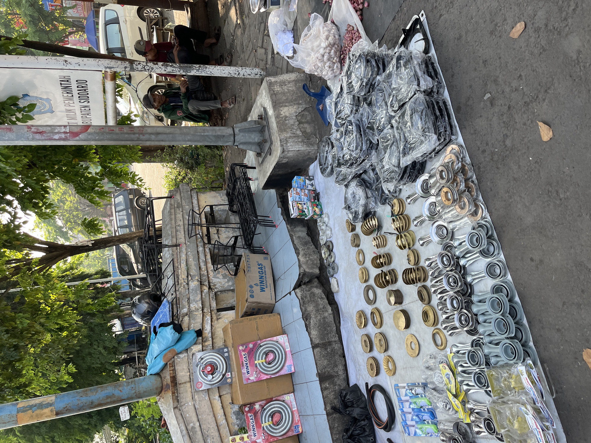 This image depicts an outdoor market stall in Juwet Kenongo, Indonesia, likely situated along a roadside or public area. The stall is displaying a variety of items for sale, predominantly kitchenware and related accessories. In the foreground, a large assortment of stove burner parts, including brass and silver-toned components, are laid out on a white tarp. Several packages of what appear to be gas stove hoses and regulators are also visible. Behind these, stacks of bagged stove burner assemblies are neatly arranged. Further back, on a raised concrete ledge, are boxed gas stove regulators with "WINNGAS" branding, as well as loose burner grates and other metal kitchen equipment. A blue jacket and a black helmet are casually placed on the ledge. In the background, two men are seated on a wooden bench, possibly vendors or customers, engaged in conversation or using their phones. A sign for the "PEMERINTAH KABUPATEN SIDOARJO" (Government of Sidoarjo Regency) is visible on a pole, indicating the administrative region. A white van and a dark SUV are parked nearby, suggesting a public thoroughfare. The scene is bathed in natural sunlight, with trees providing shade in some areas. The overall impression is of a busy marketplace selling essential household goods.