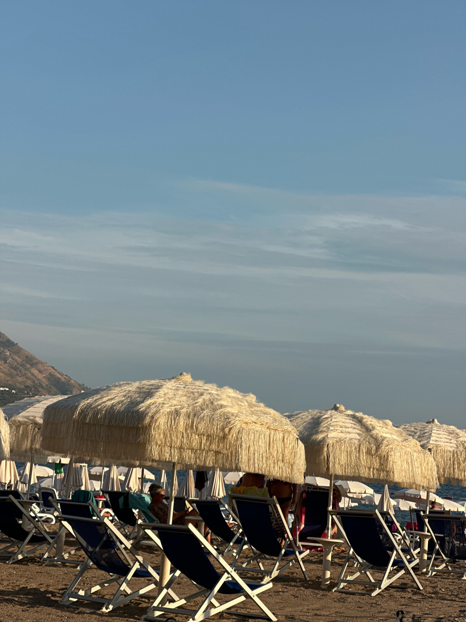 The image depicts a beach scene with rows of empty, folded chairs and large, thatched umbrellas. In the foreground, several blue canvas beach chairs with white frames are visible on a sandy surface. Behind these, more chairs and umbrellas are arranged, with some people seated under the umbrellas. The umbrellas are made of natural straw and have a fringed edge. In the background, the blue ocean and a hazy coastline with a mountain are visible under a pale blue sky with wispy clouds. The lighting suggests it is late afternoon or early morning. This scene is located on the beach in Boreti, Montenegro.