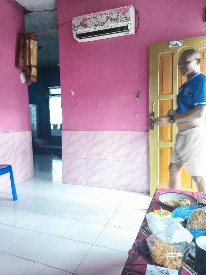 The image shows the interior of a home in Majalengka, Indonesia. The room has pink walls and white tiled floors. There is an air conditioner mounted on the wall, a blue plastic chair in the corner, and a partially obscured doorway leading to another room. A man is standing in the doorway, partially obscured from view, wearing a blue shirt and beige pants. A table sits in the foreground with assorted snacks and dishes on it.