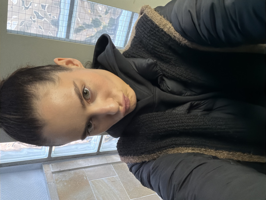 A female subject, appearing to be a young adult, is depicted in a close-up selfie taken from a high angle. Her dark hair is pulled back into a bun. She has a pouting expression with pursed lips and light-colored eyes. She is wearing a black hooded sweatshirt underneath a black puffer jacket, over which she has a dark, textured knitted vest or shawl with lighter brown trim. The background on the left shows a light-colored, possibly tiled or stone wall. To the right, a large window composed of multiple square glass blocks, some textured or frosted, allows light into the indoor setting. The specific location within the city of Unknown, Unknown is an interior space, possibly a hallway or stairwell, characterized by the glass block window and stone wall.