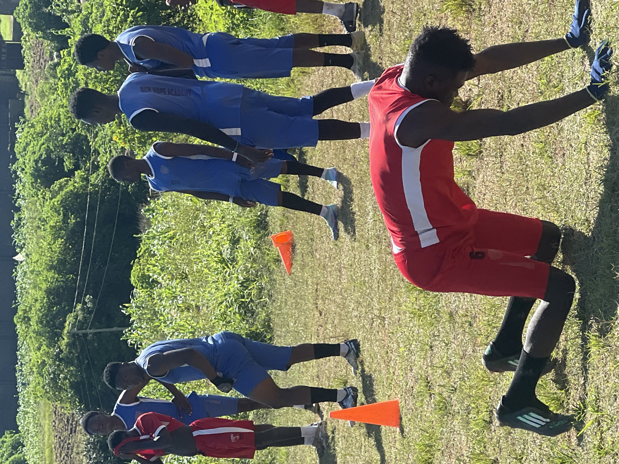 The image depicts a group of young men, likely athletes, training outdoors on a grassy field. The location appears to be within the city of Jos, Nigeria, suggested by the context and environment. In the foreground, a young man in a red athletic uniform is performing an exercise, possibly a burpee or a plank, with his hands on the ground and his body in a kneeling position. He is wearing black socks and cleats.
Several other young men are visible in the background, also dressed in athletic attire. Some are wearing blue sleeveless jerseys and blue shorts, while others are in red jerseys and shorts. There are orange training cones placed on the field, indicating a drill or practice session is in progress. The background shows lush green vegetation and a hint of a rural or semi-rural landscape with some structures in the distance. The overall scene suggests a sports training activity taking place in an open, natural environment.