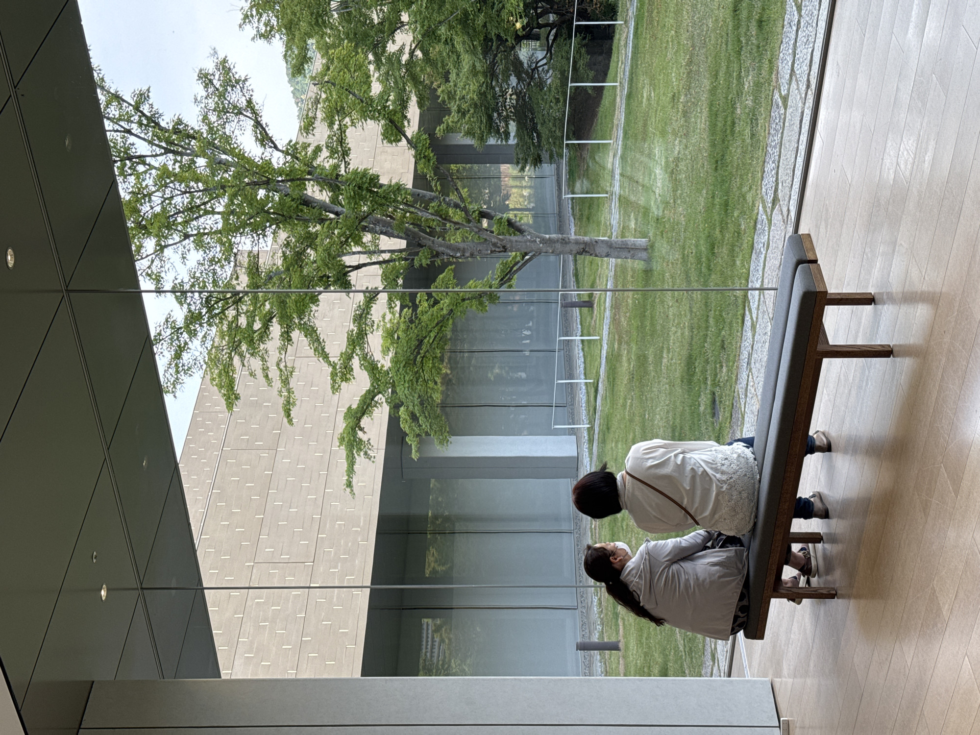 Two women are seated on a dark grey bench in an indoor space with wood flooring, observing a view through a large glass window. Outside, a lush green lawn is visible, along with a tree and a building with a textured grey facade. The building's exterior is reflected in the glass. One woman is wearing a white shirt and the other is in a light-colored jacket and mask. The setting is the Kyoto City KYOCERA Museum of Art in Kyoto, Japan.