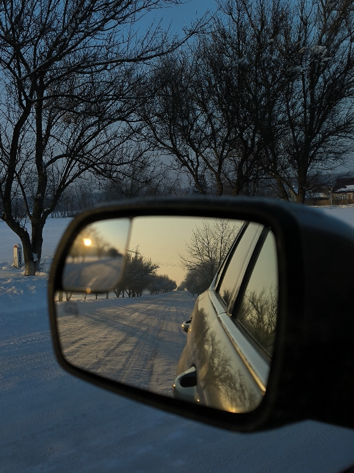 The image presents a serene winter landscape viewed from the interior of a vehicle, predominantly through its side mirror. The main subject is the car's right-hand side mirror, which reflects a snow-covered, straight road stretching into the distance. In the mirror's reflection, the sun, positioned low on the horizon, emits a bright, golden glow, casting long shadows across the pristine white snow. A line of bare, dormant trees flanks both sides of the reflected road, their branches appearing delicate against the warm light. The side of the car itself is also visible in the mirror, reflecting the same warm, winter sky and trees.
Outside the mirror's frame, directly in front and to the left of the car, are numerous large, bare trees, their intricate branches reaching towards a soft, blue sky. These trees appear dark and silhouetted against the lighter sky, with hints of snow or frost clinging to their branches. The ground outside the car is entirely covered in a thick blanket of snow. To the far left, a white post or marker stands in the snow near the road, with some faint, unreadable text or markings on its surface.
The activity suggests a quiet journey or stop along a snowy, rural road during either sunrise or sunset, indicated by the low angle and warm color of the sun. The setting is a cold, wintry environment in Khrinivka, Ukraine, characterized by snow and dormant vegetation. The mood evoked is one of peaceful solitude and the quiet beauty of winter.