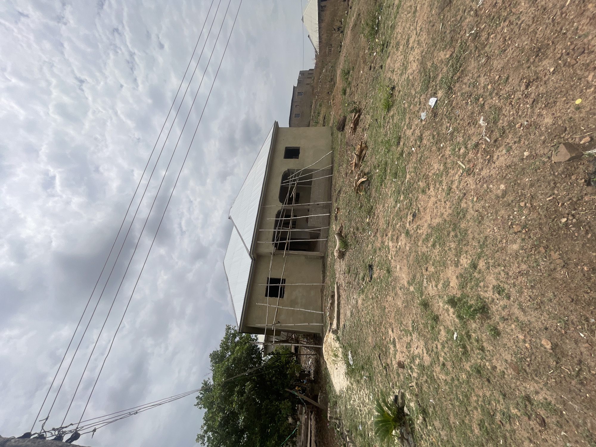The image shows a partially constructed building in Lokoja, Nigeria. The building appears to be a two-story structure with a white corrugated metal roof. Bamboo scaffolding is erected around the exterior walls. An open window is visible on the upper level. A power line is positioned in the foreground with four visible electrical cables. The surrounding environment is characterized by sparse vegetation, brown soil, and scattered debris. A tree is visible in the left foreground, partially obscuring the base of the building. In the background, another building is partially visible on a slight incline. The sky is filled with overcast clouds.