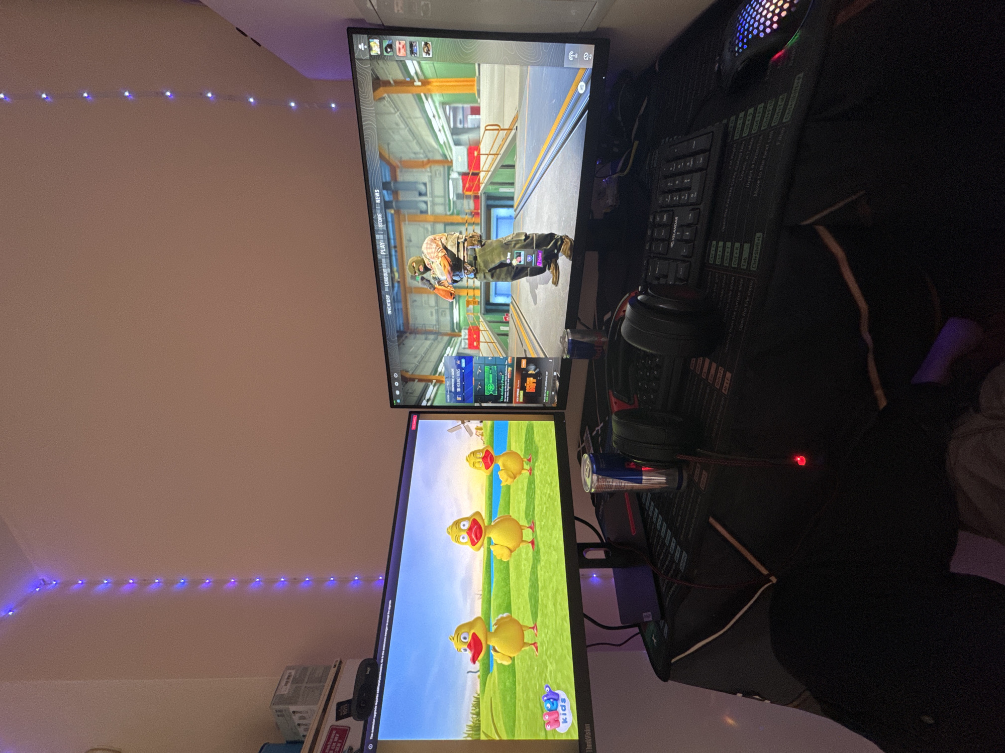 The image shows a desk setup with two computer monitors. The top monitor displays a first-person shooter video game, and the bottom monitor shows a children's cartoon with animated ducks. A keyboard and mouse are on the desk, along with headphones and cans of Red Bull. The environment is a room with blue LED lights along the wall and ceiling. It is not possible to determine the location in Sofia, Bulgaria from this image.