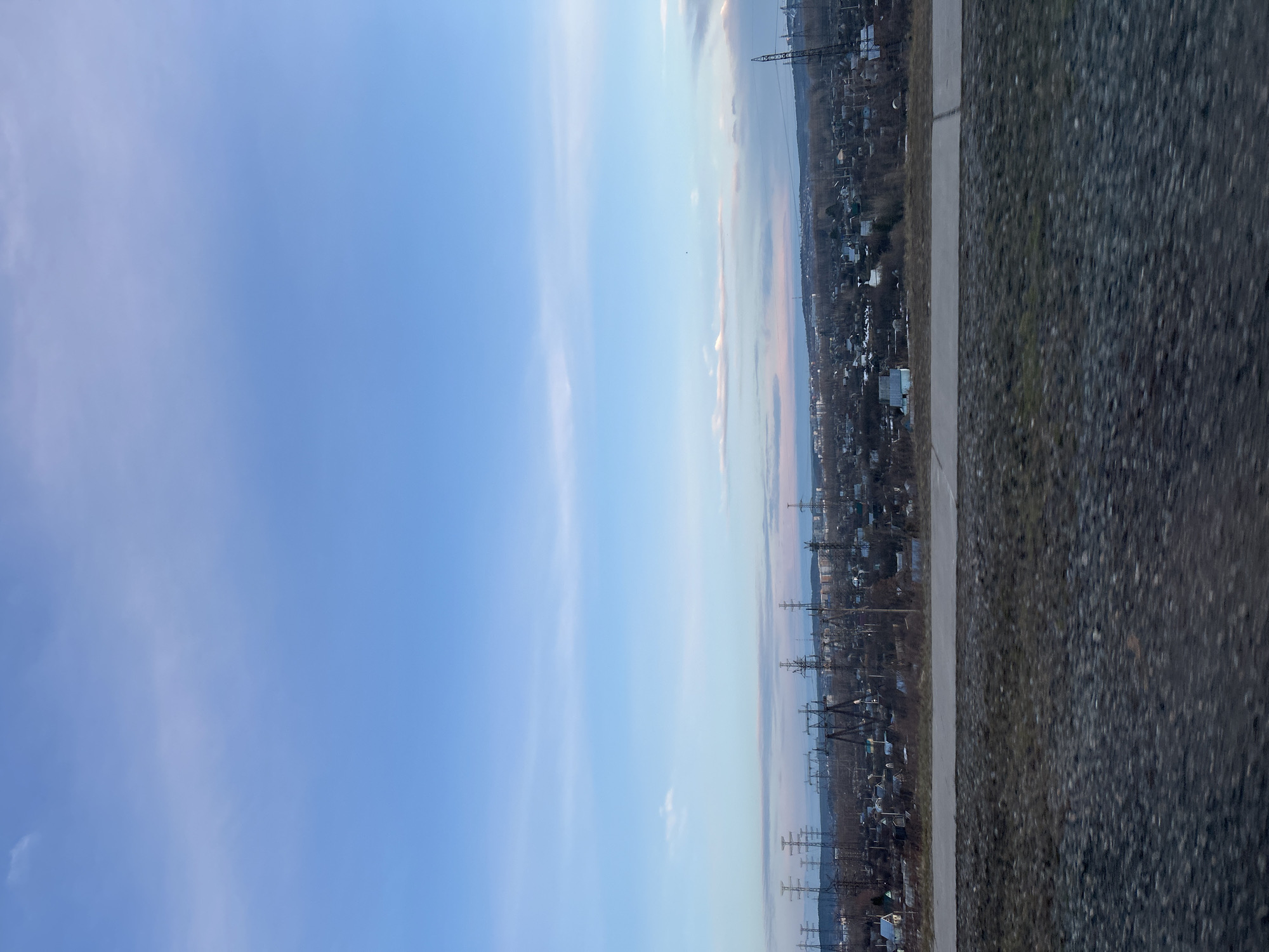 The image shows a view overlooking a section of Chaikovsky, Russia. Power lines run through the landscape, and residential buildings can be seen in the distance. A portion of asphalt or pavement takes up the right side of the frame. The sky is predominantly blue with some scattered clouds.