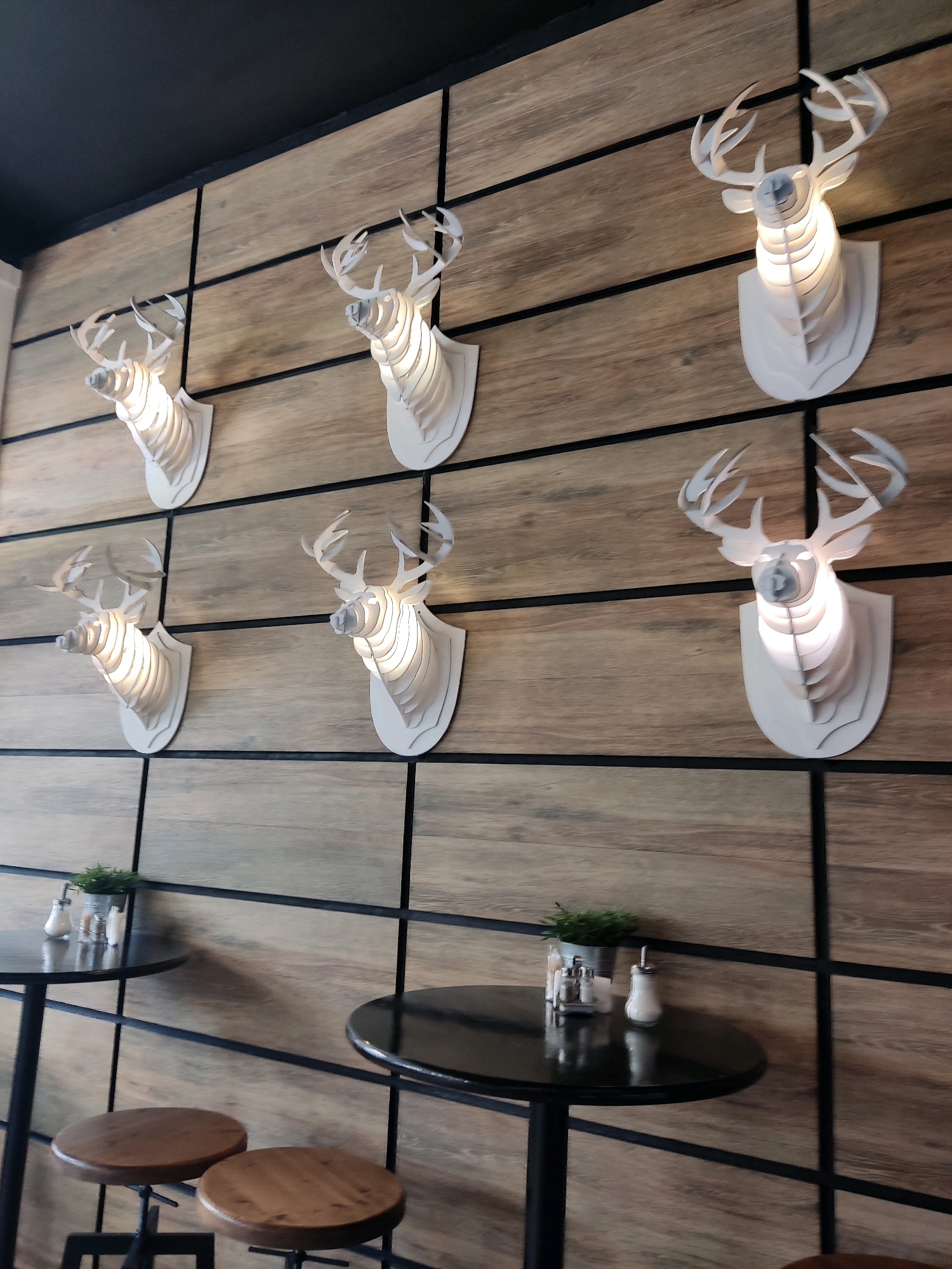 The image depicts an interior scene with a rustic-modern aesthetic, featuring a wall decorated with six white, stylized deer head sculptures. These sculptures are illuminated from within, casting a warm glow and creating a layered effect. The wall itself is paneled with wood planks arranged horizontally, separated by vertical black metal strips. In the foreground, two black round tables are visible, each accompanied by wooden stools. One table is set with a small potted plant and salt and pepper shakers, while the other has fewer visible condiments. The overall atmosphere suggests a contemporary dining or lounge space. There are no people or animals present in the image. The scene is static and indoors.
