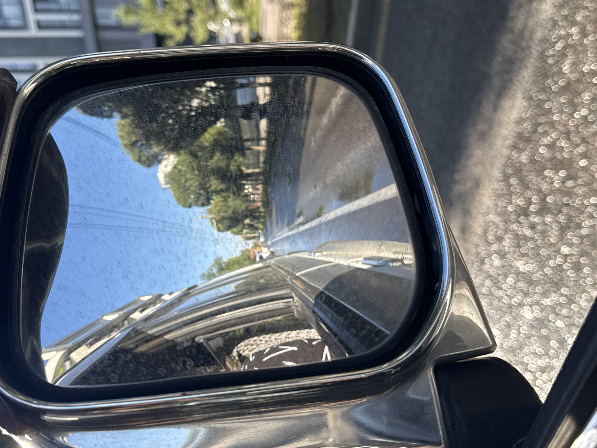 The image shows a side-view mirror of a vehicle, reflecting a street scene in Almaty, Kazakhstan. The mirror's surface has water droplets, creating a blurred effect. Through the mirror, a road with a white line is visible, along with trees and buildings on the side. The sky is blue. The vehicle appears to be in motion, as suggested by the reflection of the road and the slight blur. The foreground shows the exterior of the vehicle, including the mirror housing and part of the car's body. The ground beneath the vehicle is covered in specular highlights, likely from sunlight reflecting off small water droplets or textured surfaces. The reflection in the mirror shows a street with parked cars and some pedestrian activity in the distance. One can see a building with multiple stories and balconies on the right side of the reflection. The general environment suggests a sunny day.