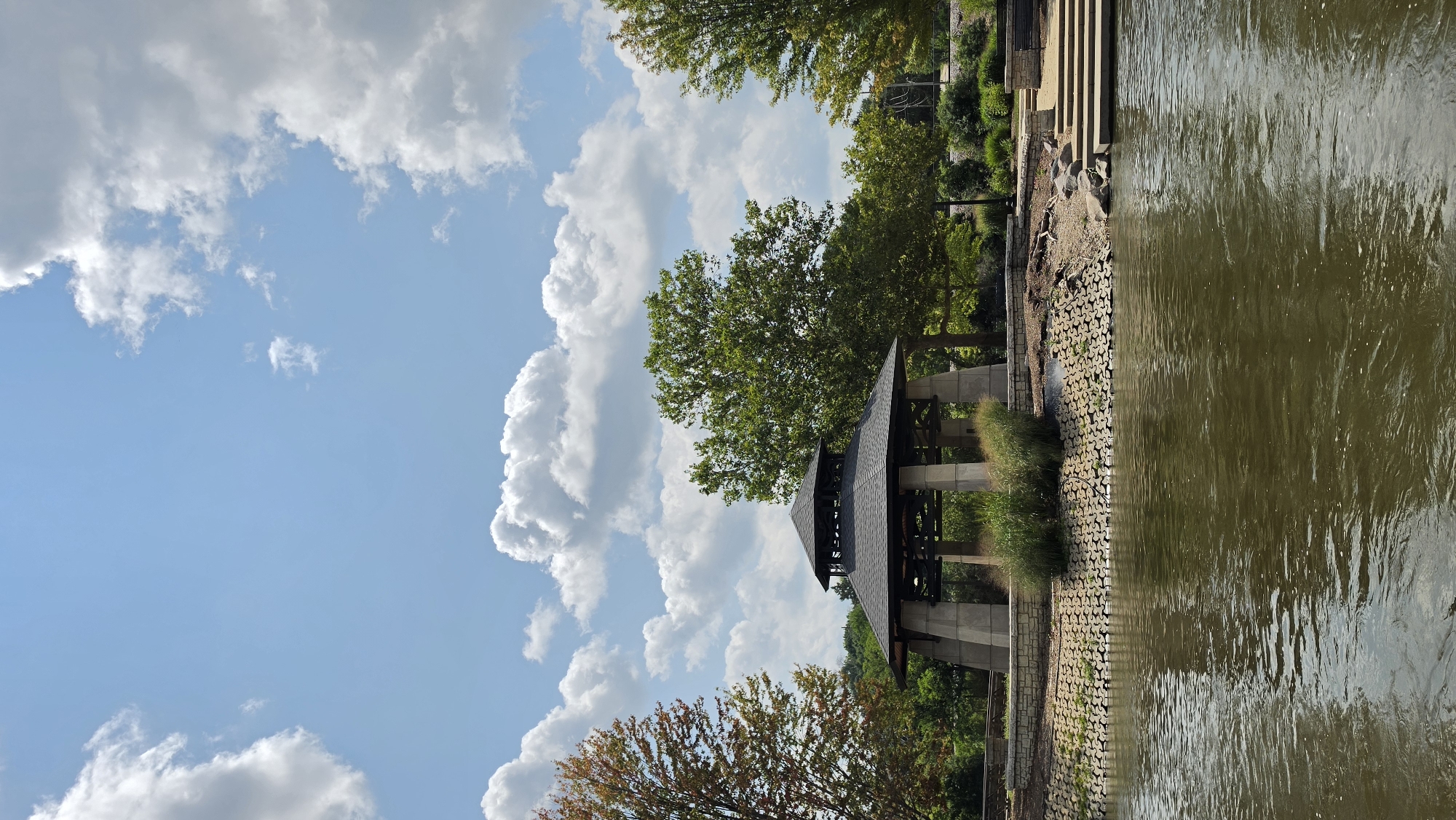 This is a landscape photograph of a gazebo and riverbank, likely taken from the water. The image depicts a dark, multi-tiered gazebo with a peaked roof supported by stone pillars. The gazebo is situated on a raised, stone-lined embankment, with ornamental grasses and shrubs growing around its base. To the left of the gazebo, a tree with autumnal foliage is visible, while a lush green tree stands to its right. The river in the foreground is a murky brown, with ripples on its surface reflecting the sky. The riverbank is fortified with a textured, interlocking material and features a set of concrete steps leading up to a paved area with benches. The background reveals more trees and vegetation, with a clear blue sky dotted with large, white cumulus clouds. The scene is set in a park-like environment, potentially along a riverfront in the city of Elgin, United States of America. There are no people visible in the image.
