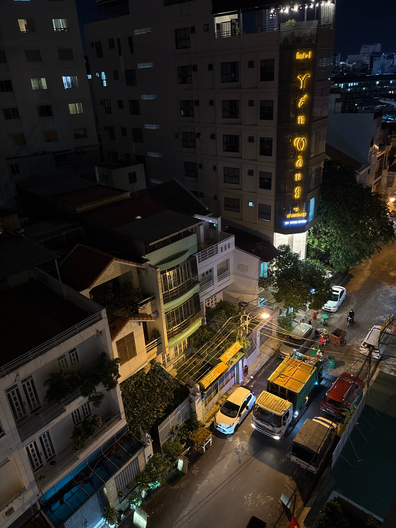 This aerial nighttime photograph depicts a street scene in Nha Trang, Vietnam. The image captures several multi-story buildings, including a prominent hotel with illuminated yellow lettering spelling out "YEN YANG". A narrow street is visible, with several vehicles present, including a garbage truck, a white car, a red car, and a white van. A person is seen riding a motorcycle, and a few other individuals are standing near the garbage truck. Power lines crisscross above the street, and trees are interspersed between the buildings. The overall impression is one of an urban environment at night, with streetlights and building lights illuminating the scene.