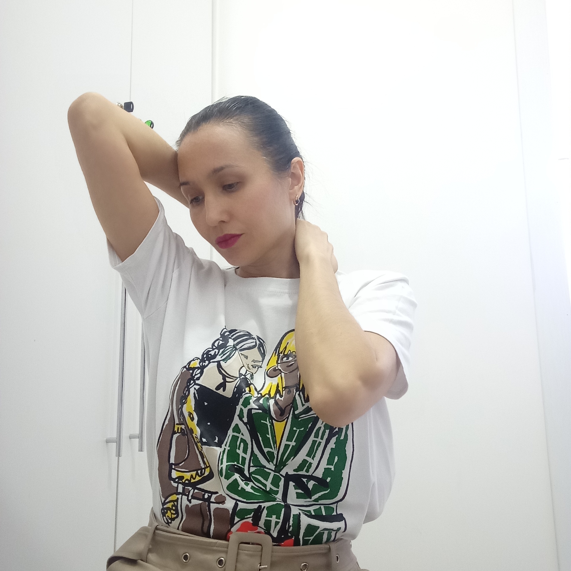 A woman is posing indoors. She has dark hair pulled back, red lipstick, and is wearing a white t-shirt with a cartoon graphic and a beige belt. Her arms are raised, with one hand behind her head and the other touching her neck. The background consists of white walls and a white cabinet with silver handles. There is nothing in the image to indicate that it was taken in Izhevsk, Russia.