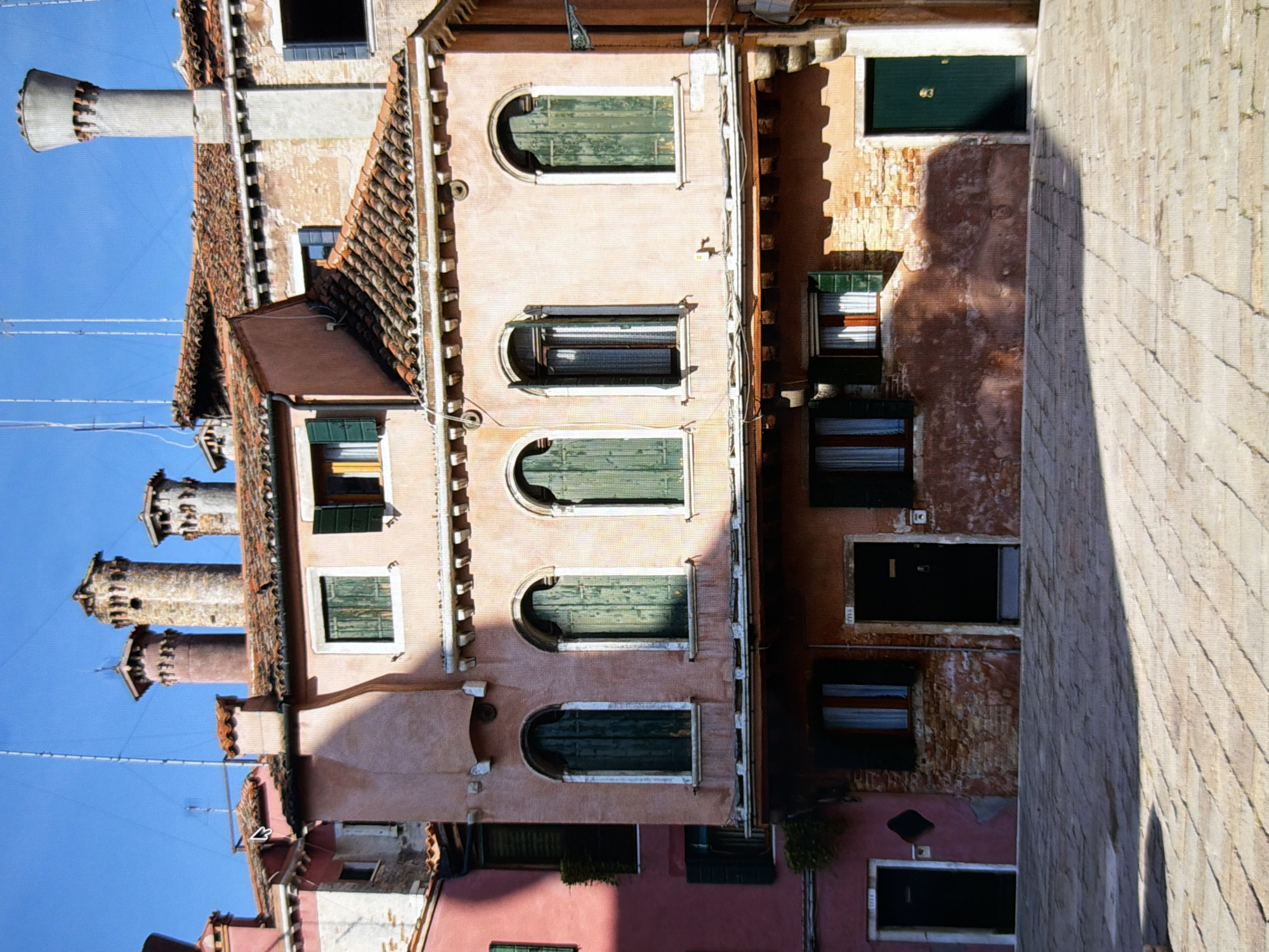 The image depicts a narrow cobblestone alleyway in Venice, Italy, bathed in bright sunlight. The scene is dominated by the facades of old, pastel-colored buildings with terracotta tiled roofs. Several windows with green shutters are visible, some open and some closed. One building features arched windows with green wooden shutters. In the foreground, the textured pattern of the cobblestones is clear, with shadows casting across the ground. The overall impression is one of historic architecture and a tranquil urban setting. There are no people visible in the image. The architecture suggests a European city, specifically Venice, with its distinctive building styles and materials.