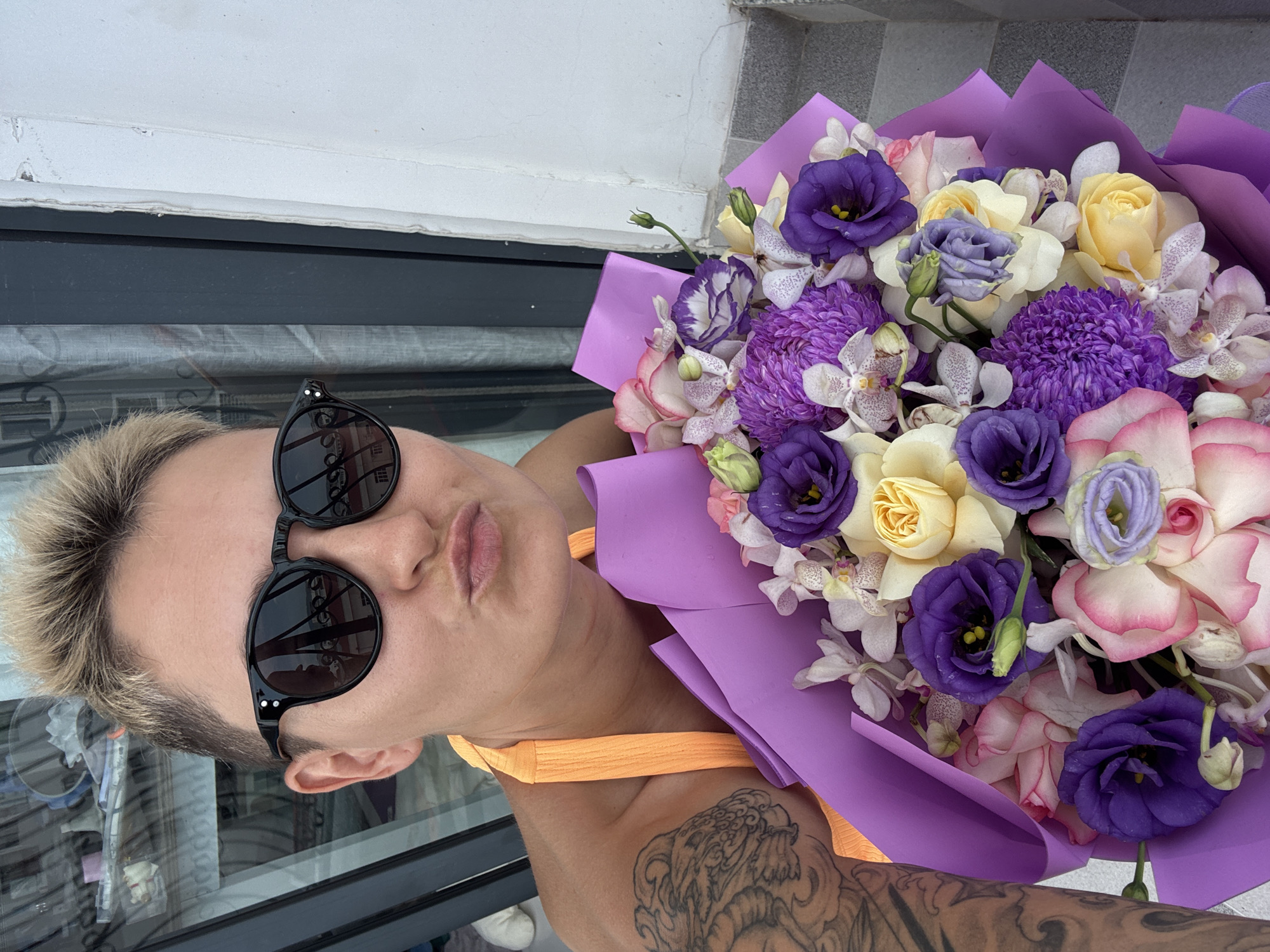 An unidentifiable person with short, spiky blonde hair, wearing black sunglasses and an orange halter top, is holding a bouquet of purple, pink, and yellow flowers wrapped in purple paper. The person is puckering their lips and looking towards the camera. A large tattoo is visible on their left arm. The background shows a window with reflections of buildings and what appears to be a balcony railing. The scene is outdoors, with a wall and tiled floor visible on the right side of the image. The setting appears to be a balcony or patio overlooking an urban area, possibly in Nha Trang, Vietnam, given the presence of buildings and the general architectural style.