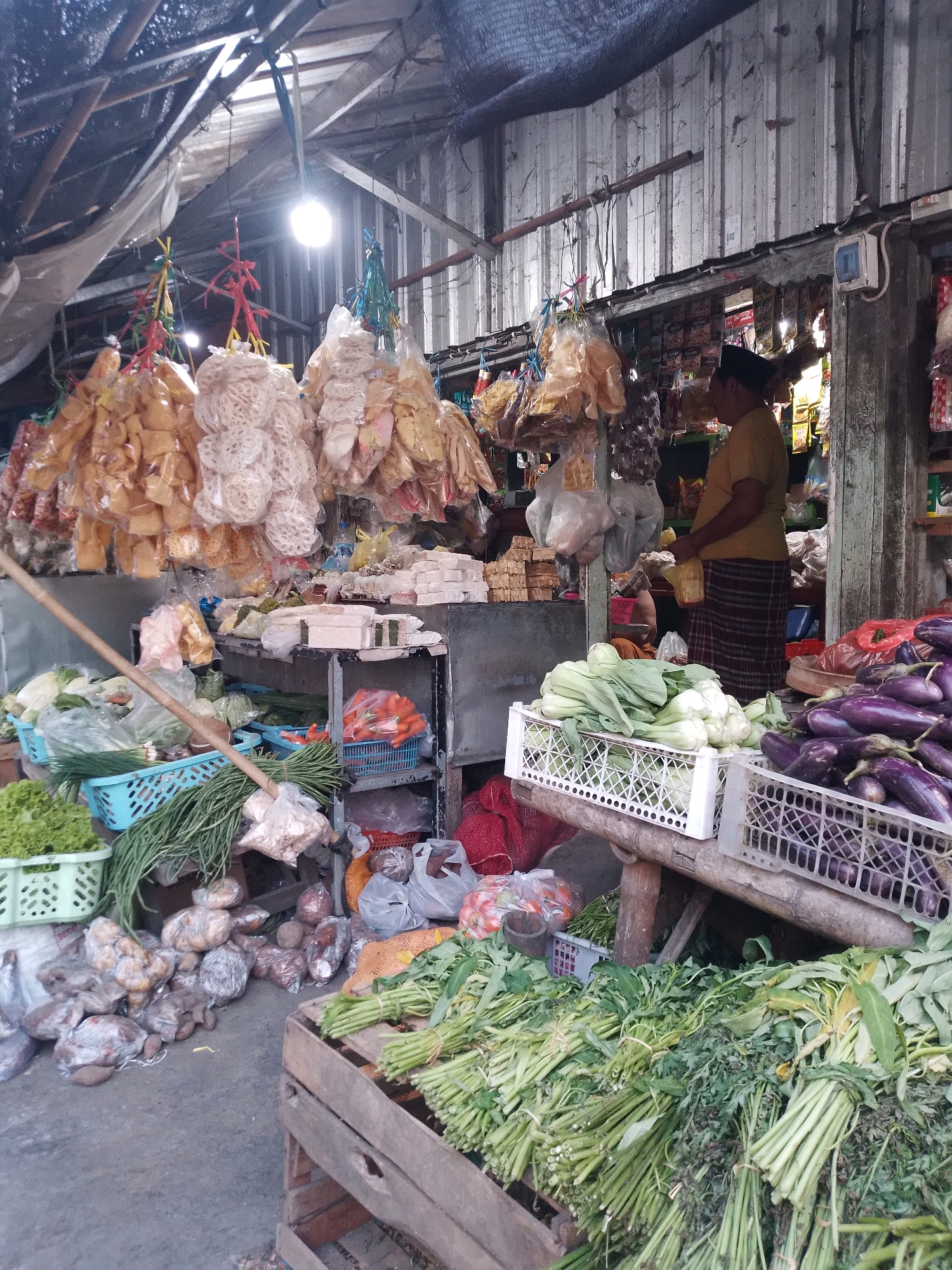 The image depicts an outdoor market scene in Kertosono, Indonesia, likely a fresh produce and snack market. A vendor, a person of indeterminate gender wearing a yellow shirt and a patterned sarong, stands near a stall in the background, holding a bottle. The market stall is laden with various items. In the foreground, a wooden crate is filled with bundles of green leafy vegetables, possibly water spinach or similar greens. To the left of the crate, more green vegetables and some vegetables wrapped in plastic are visible on the ground. Further into the market, blue plastic crates hold more vegetables, including lettuce, long beans, and carrots. Bags of snacks, appearing to be crackers or crisps, are hanging in plastic packaging from the ceiling. The market appears to be covered by a makeshift roof structure made of corrugated metal and wooden beams. The overall atmosphere is one of a bustling, local market.