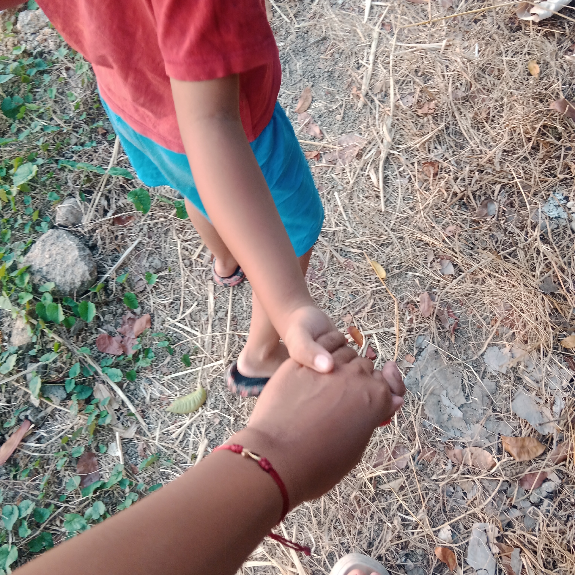 A close-up shot focuses on two hands clasped together, held by an adult and a child. The adult's hand, in the foreground, is adorned with a red string bracelet. The child's hand is smaller and appears to be that of a young boy, wearing a red t-shirt and blue shorts. Their feet are visible, with the child wearing flip-flops. The scene unfolds on a dry, earthy path, scattered with dried leaves and grass, indicative of an outdoor setting. Green foliage and a large rock are visible to the left, suggesting a natural environment. The lighting suggests it is daytime. The location is Tegowanu Kulon, Indonesia.