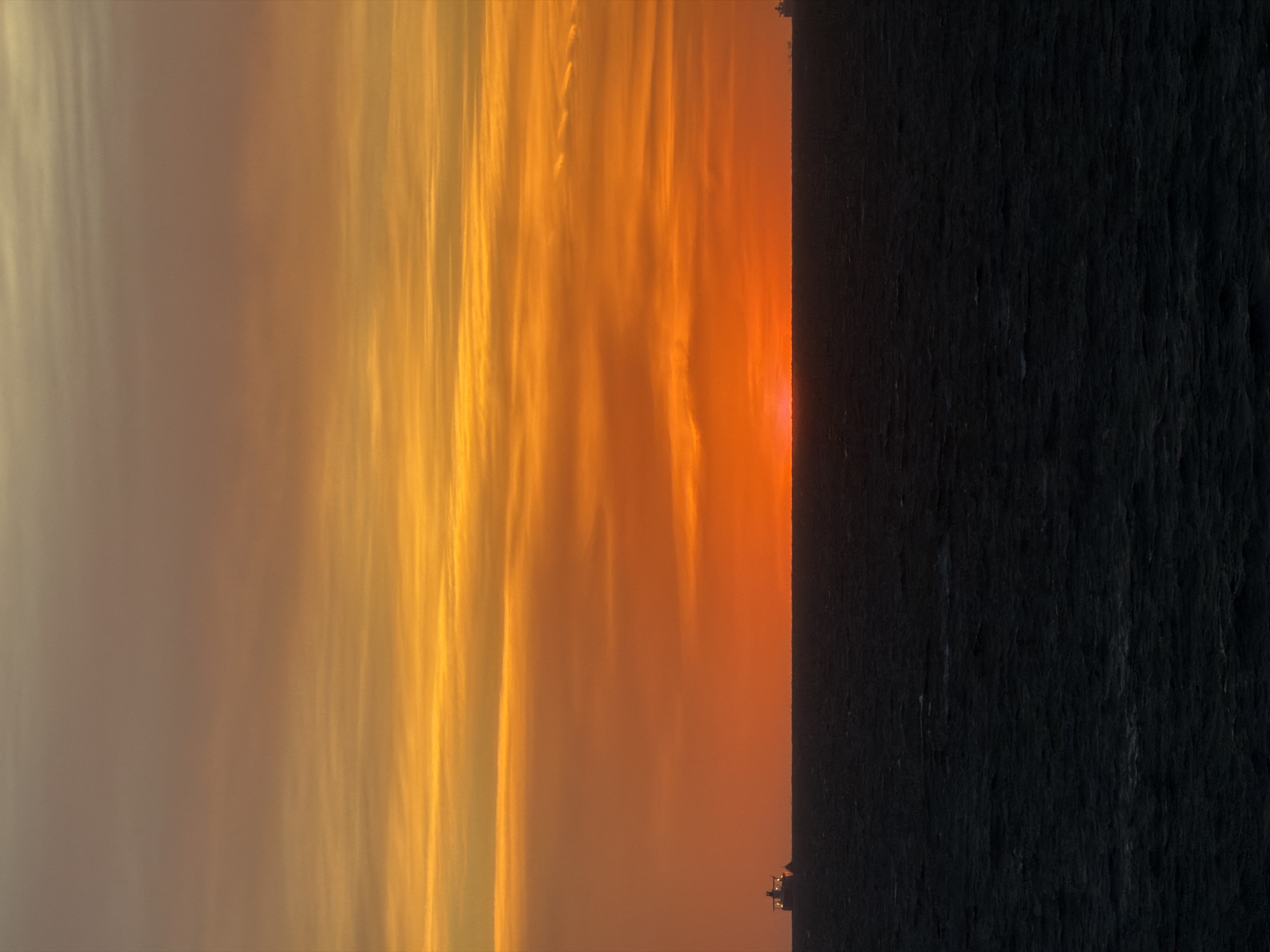 This media file presents a stunning seascape at either sunrise or sunset. The scene is dominated by a vast, vibrant sky painted in a gradient of warm, intense colors, with a dark, silhouetted body of water below.
The sky transitions dramatically from a muted, soft golden-brown on the left side of the frame to brilliant golden yellow, fiery orange, and deep red towards the right. Long, ethereal clouds, appearing as streaks and wisps, stretch across the sky, catching the intense light and glowing against the fiery backdrop. The patterns of these clouds suggest atmospheric movement and create a dynamic texture in the upper half of the image.
The sun itself is partially visible as an intense, glowing red-orange disc just peeking above the horizon on the far right, casting its last or first light across the clouds.
Below the vividly colored sky, a vast expanse of water occupies the bottom portion of the image. It appears almost black, completely silhouetted against the bright horizon, offering a stark contrast to the brilliant sky. The water's surface shows subtle textures, indicating gentle ripples or small waves, but no distinct features.
A sharp, straight horizon line clearly separates the radiant sky from the dark water. A tiny, indistinct cluster of lights or structures is barely visible in silhouette on the horizon line towards the right, possibly a distant vessel or coastal feature.
No people or identifiable objects are present apart from the natural elements and the minuscule distant structure. There is no visible text. The overall impression is one of majestic natural beauty and the breathtaking spectacle of dawn or dusk over the ocean.