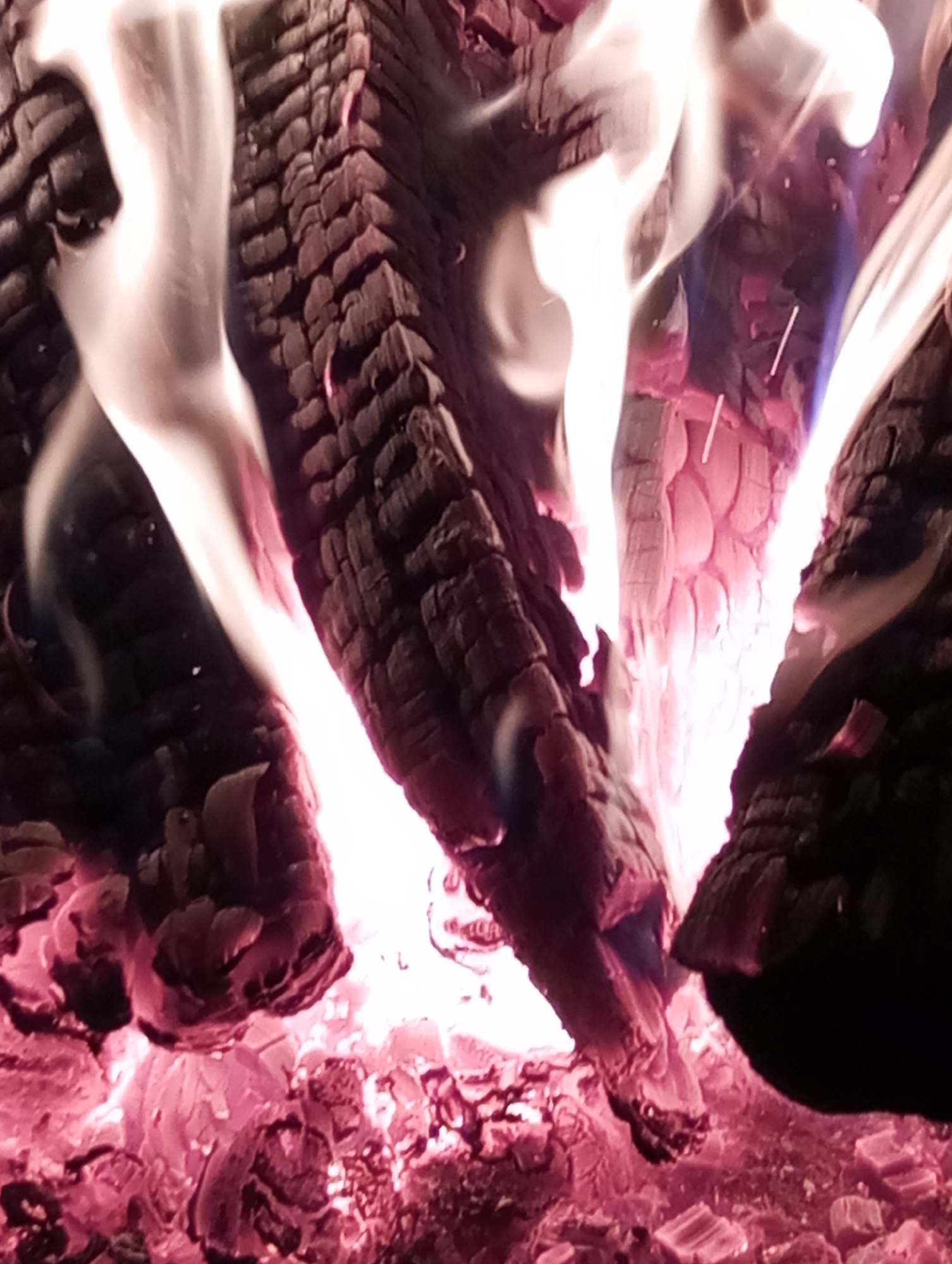 The image shows a close-up view of burning logs and embers. The logs are charred and textured, with flames rising between them. The embers glow red-hot at the base. The location in the city of Буренка, Russia is not evident from the visual information.