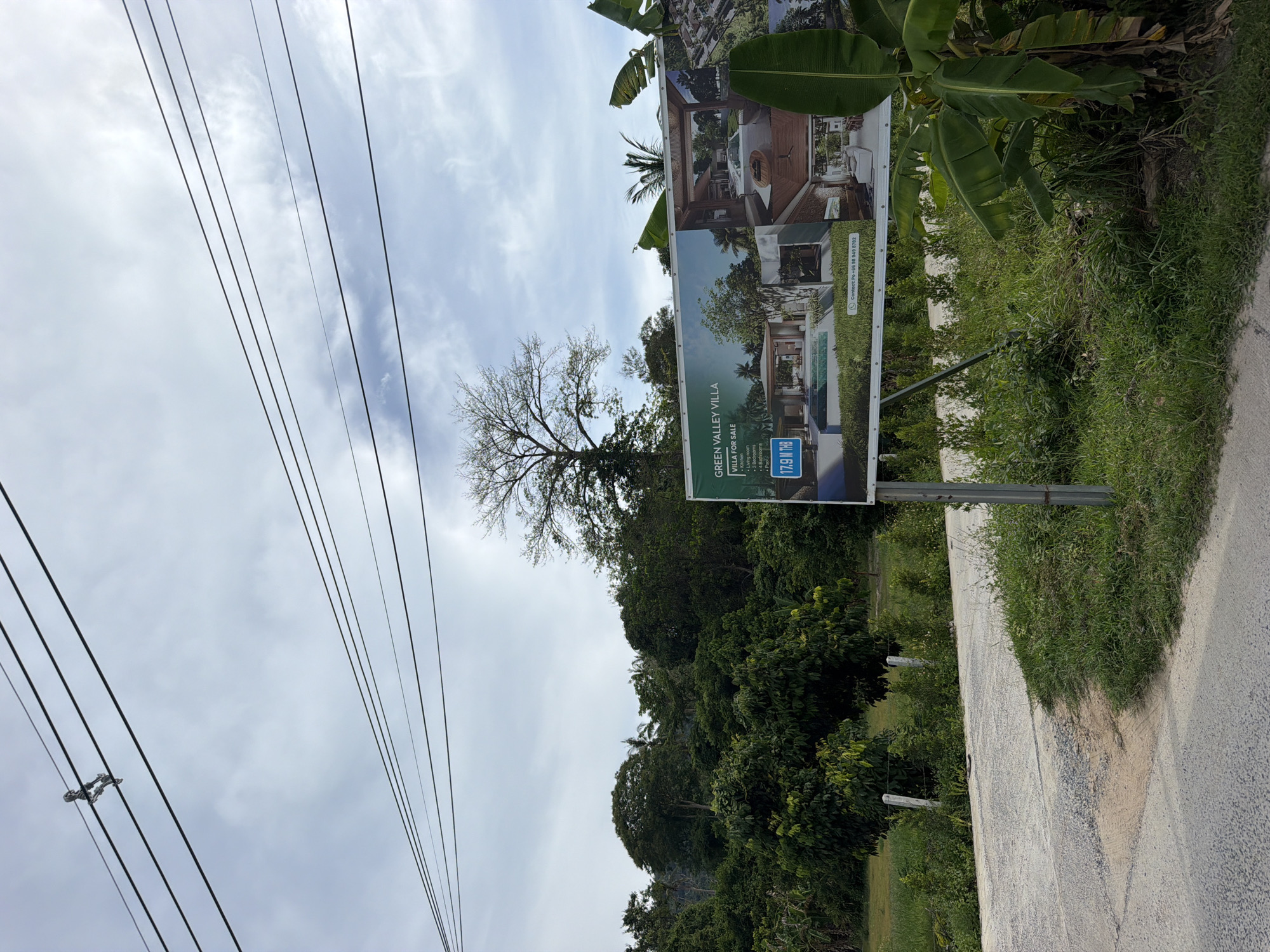 The image shows a billboard for "GREEN VALLEY VILLA VILLAS FOR SALE" placed in an outdoor setting in Baan Mae Nam, Thailand. The billboard displays images of modern villas with swimming pools and lush greenery. Text on the billboard indicates features such as a kitchen, living room, 3 bedrooms, 2 bathrooms, and a pool, with a price of "17.9 M THB". A contact number, "+66 98 949 8782", is also visible.
The billboard is mounted on metal poles and is situated next to a dirt road. The surrounding environment is characterized by dense vegetation, including trees and banana plants, and a cloudy sky. Power lines are visible in the upper portion of the image. The road appears to be in a rural or semi-rural area, with no people or vehicles present in the immediate vicinity of the billboard. The overall scene suggests a property advertisement placed in a location relevant to potential buyers or at a visible point along a roadway.