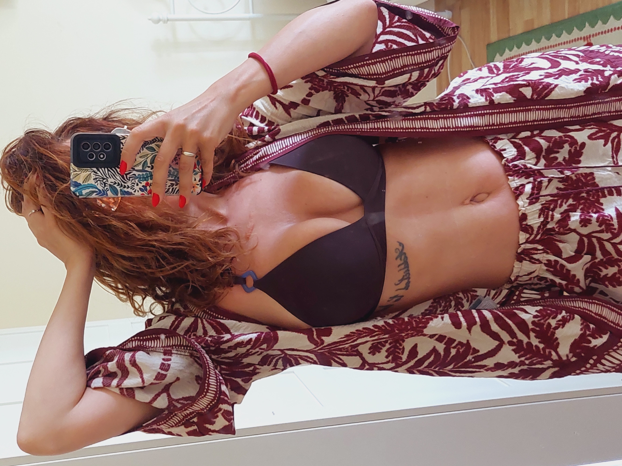 A woman with red curly hair is taking a selfie in a mirror. She is wearing a brown bikini top, a patterned robe, and matching patterned shorts. She has a tattoo on her side and is wearing rings on her fingers and a bracelet on her wrist. The room appears to be a bedroom with a white bed frame visible in the background. The image is taken in Sozopol, Bulgaria.