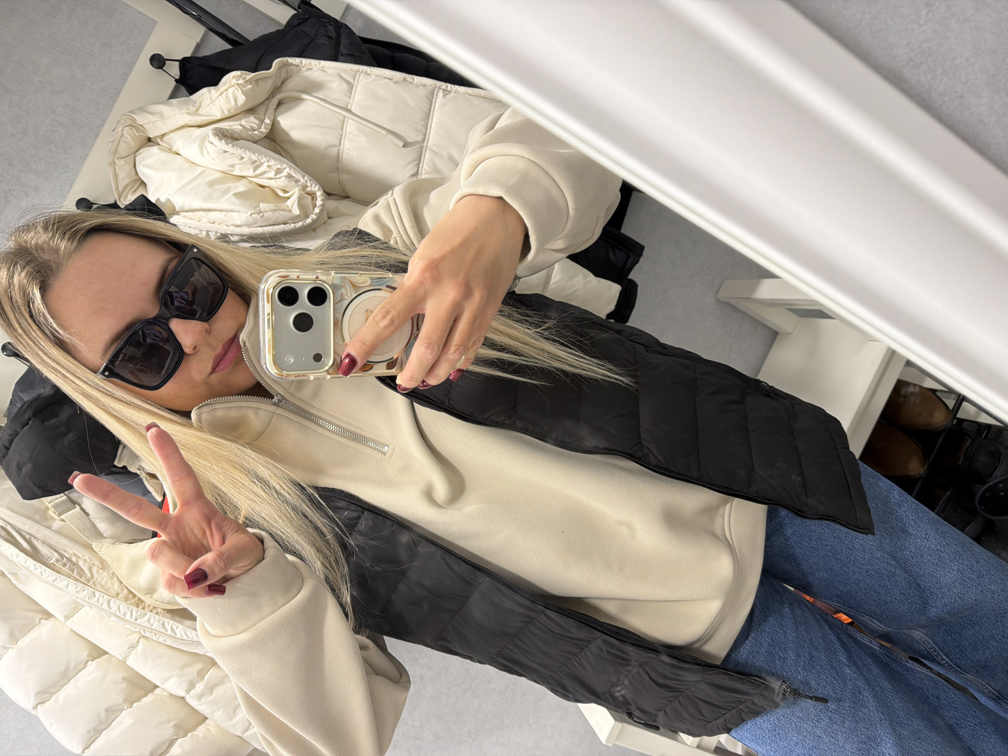 A woman is taking a selfie in what appears to be a closet or dressing area. She is wearing sunglasses, a light-colored hoodie, and a black puffer vest. She is also wearing blue jeans and has her hair down. She is holding a smartphone with a decorated case in her right hand, taking the picture, and making a peace sign with her left hand. There are several jackets and coats hanging in the background, including a white puffer jacket. The environment looks like an indoor storage space, with shelves visible in the background. There is nothing to suggest this photo was taken in Votkinsk, Russia.