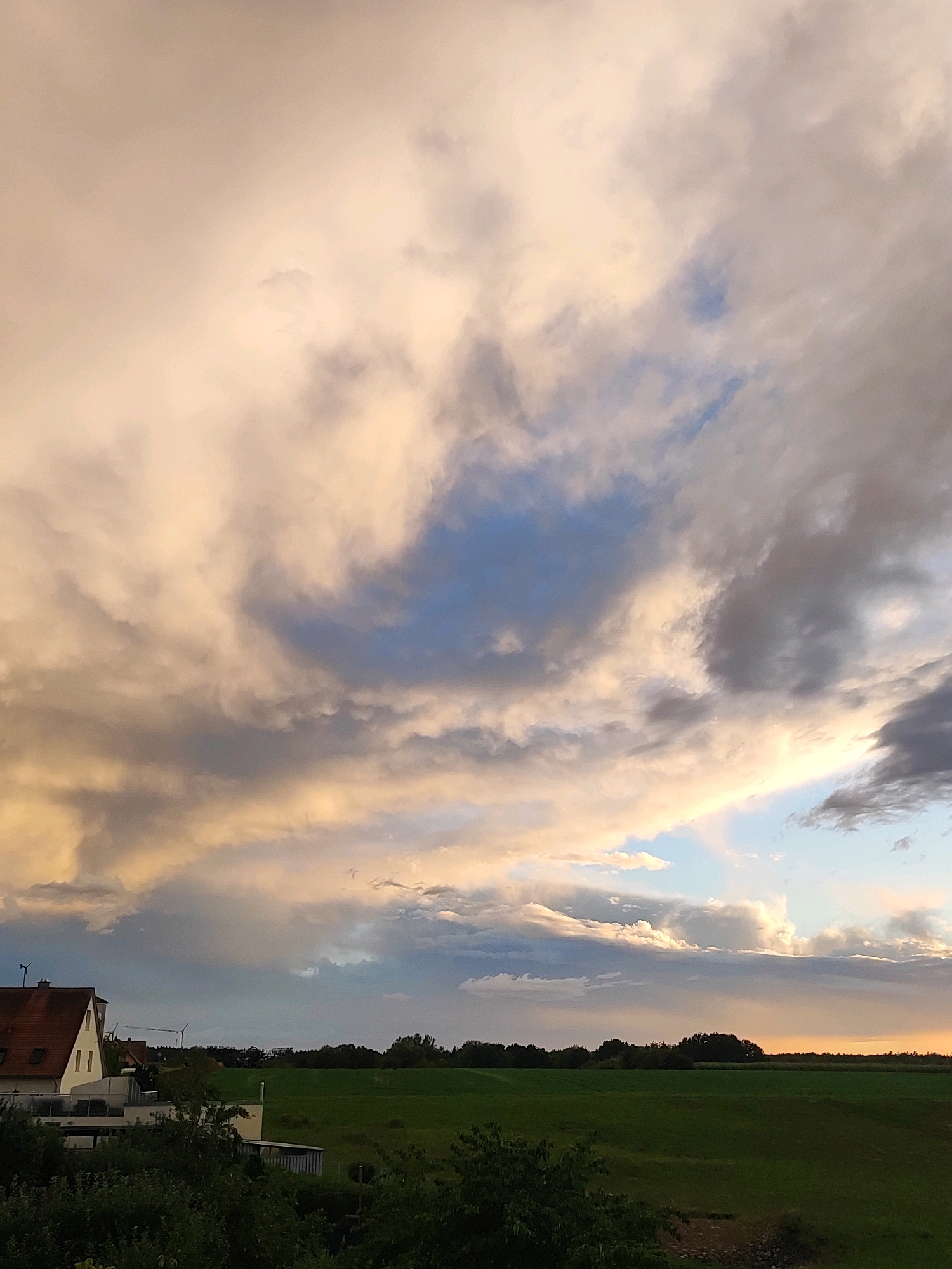 The image depicts a landscape scene in Burgoberbach, Germany. The sky is filled with dramatic, light-colored clouds, tinged with yellow and orange hues, suggesting either sunrise or sunset. Patches of blue sky are visible through the clouds. In the lower left of the image, a white building with a red roof is partially visible, characteristic of German architecture. Adjacent to it is a modern-looking building with flat roofing. Beyond these structures, a flat green field stretches towards a tree line that borders the horizon. In the distance, a construction crane is visible, indicating ongoing development or infrastructure work. The overall environment appears to be rural or semi-rural. There are no people or animals visible in the frame.