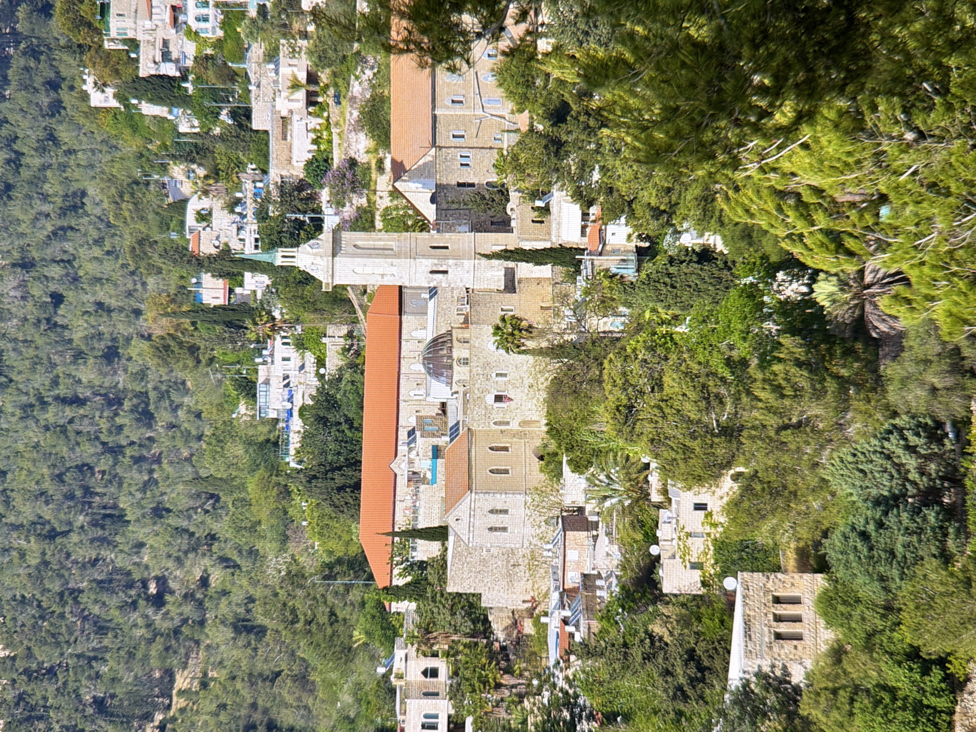 This is a high-angle, long shot, taken during the daytime, of a mountainous, verdant landscape dotted with buildings. The image is oriented vertically, with the trees and buildings appearing upside down.
The primary subjects are a collection of buildings with red-tiled roofs and stone facades, nestled into a hillside covered with dense green trees and vegetation. One prominent building has a tall, slender tower with arched windows. Another building features a dome. There are smaller structures with white walls and windows scattered throughout the scene. The architecture suggests a historical or religious site, possibly a monastery or an old town.
The scene is lush and appears to be in a rural or semi-rural setting, far from a bustling city. The trees are evergreen and appear healthy, indicating good weather. The overall impression is one of tranquility and historical significance, set against a natural backdrop. There is no text visible in the image. The orientation of the image makes it difficult to definitively assess the activity or precise emotions conveyed, but the natural setting and architecture evoke a sense of peace.