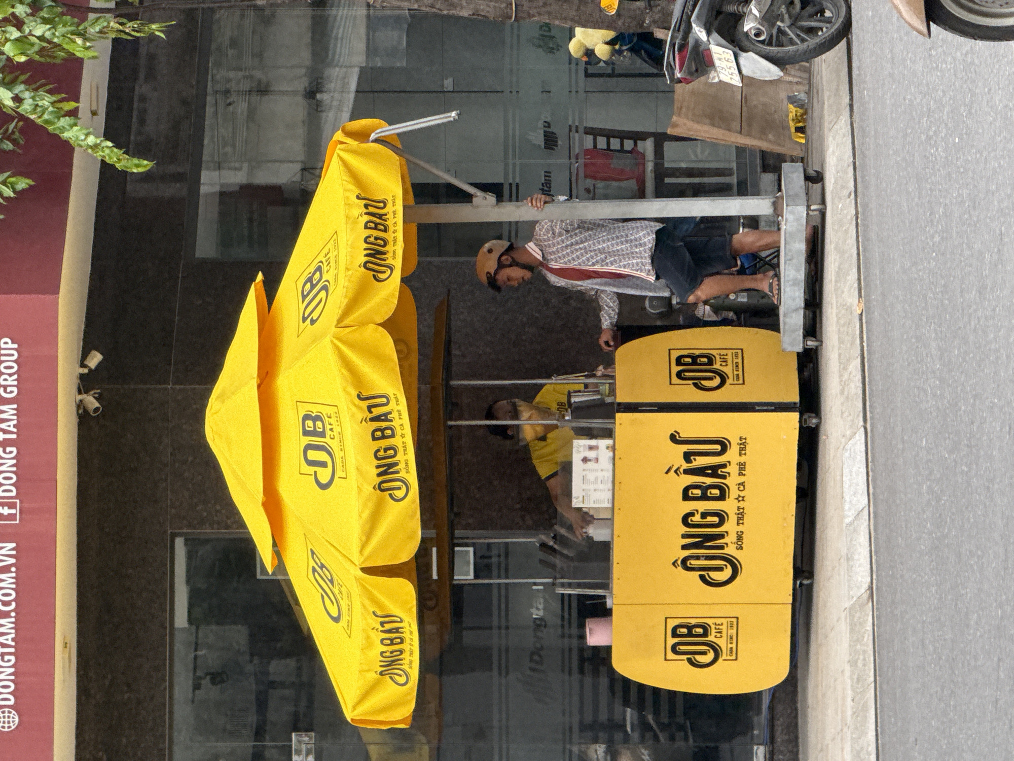 The image depicts a street scene in Nha Trang, Vietnam, focusing on a yellow coffee cart. The cart is branded with the logo "DB CAFE" and the Vietnamese text "ÔNG BẦU," meaning "Mr. Boss." A man wearing a patterned shirt, dark shorts, and a helmet stands next to the cart, holding onto its central pole. Another person, wearing a yellow t-shirt and a mask, is visible behind the counter, seemingly preparing a drink. A motorcycle is parked to the right of the cart. The background features a building with large windows, reflecting the surrounding environment. The overall impression is that of a typical urban commercial street in Vietnam.