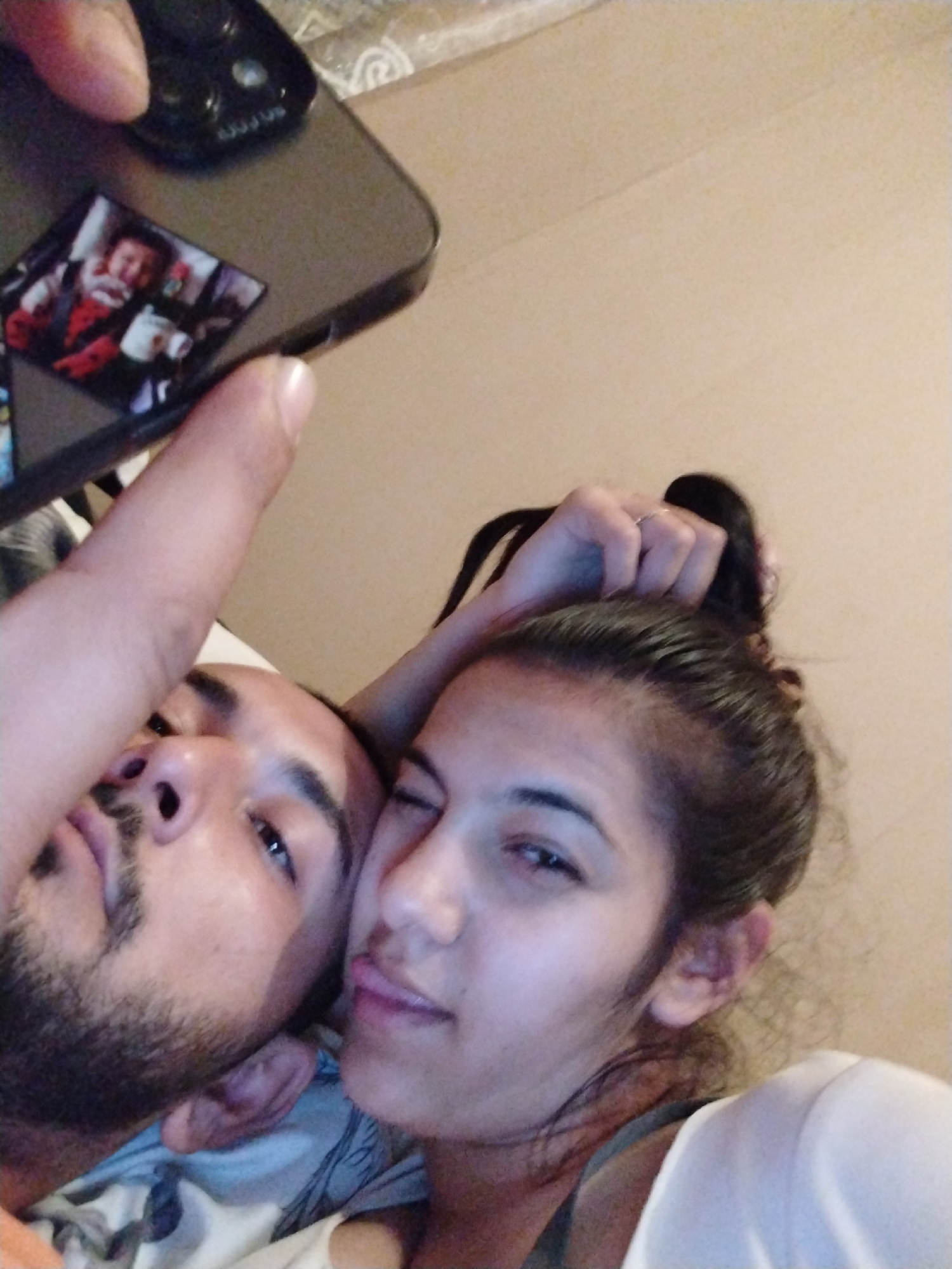 A man and a woman are taking a selfie in an indoor environment. The man has short dark hair and a beard, while the woman has her hair pulled back in a bun. She is sticking her tongue out slightly. A smartphone is held above them, with a sticker on the back. The background consists of a plain wall. The location and city are unknown.