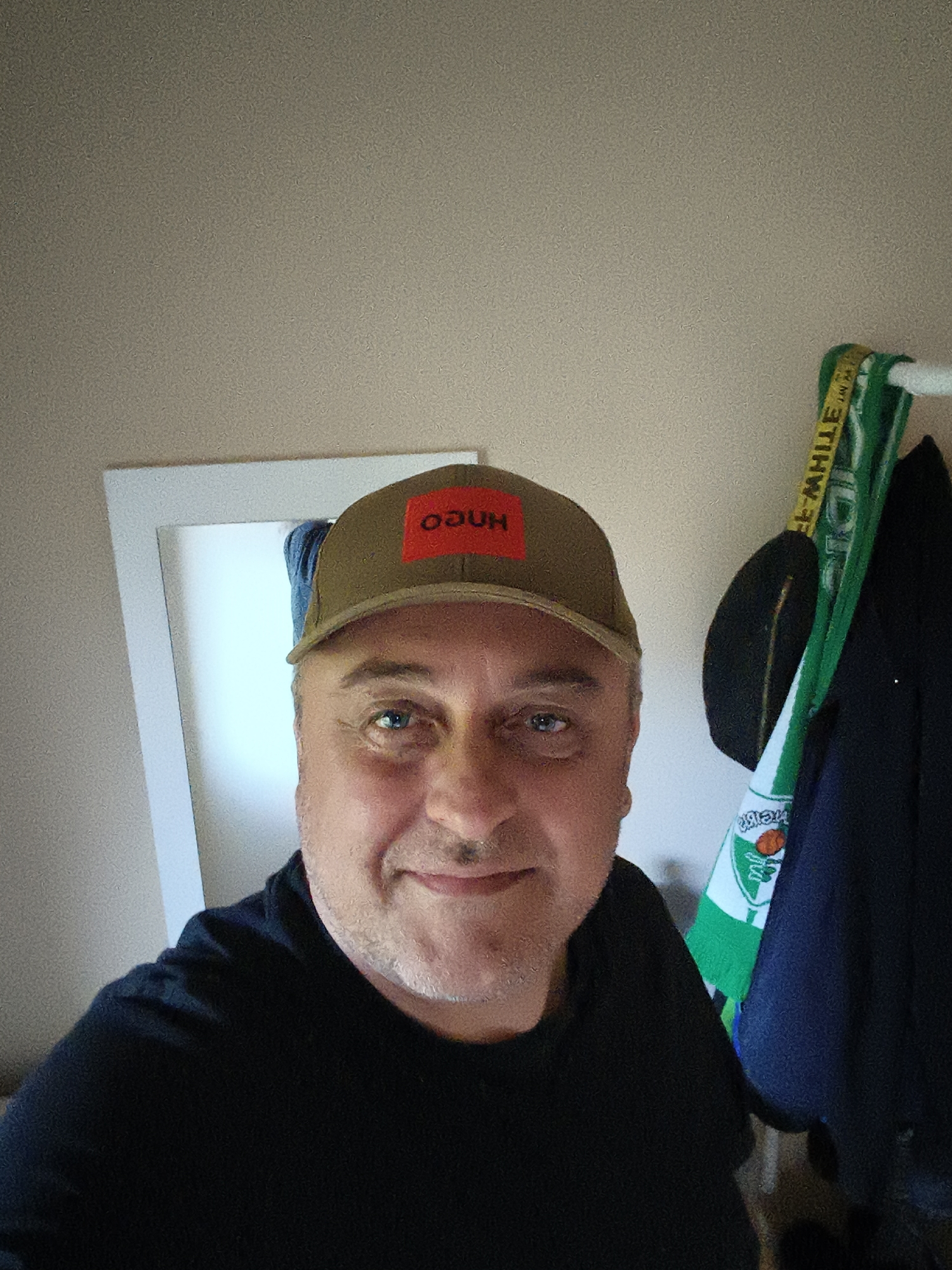 A middle-aged man with light skin, blue eyes, and light facial stubble is depicted in a selfie, smiling directly at the camera. He is wearing a black t-shirt and an olive green baseball cap. The cap features a red rectangular patch on the front, with the white text "OGUH", which is a mirrored or inverted version of "HUGO".
The setting appears to be indoors, against a plain, light beige-grey wall. To the left of the man, a portion of a white-framed mirror is visible, reflecting a light blue object. To his right, several items are hanging on a rack or hooks. These include a yellow strap or belt with partially visible black text that reads "OFF-WHITE™ IN M", a green and white striped scarf (possibly a sports scarf with an indistinct logo), and a dark blue garment, likely a jacket or coat. The man's expression is friendly and content. The lighting is soft and even, typical of an indoor environment.