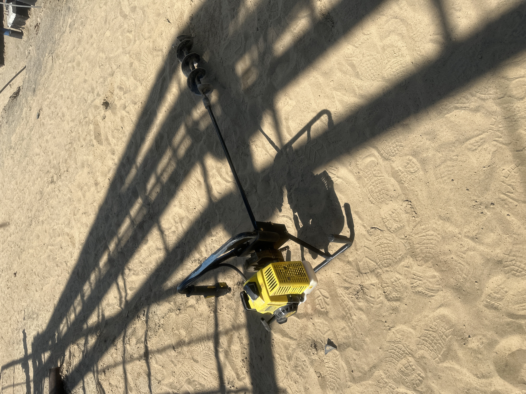 An auger is lying on the sandy ground in Чикшино, Russia. The auger is yellow and black and has a long, spiraled drill bit attached to it. The ground is covered in sand with numerous footprints visible. Long shadows are cast across the sand, indicating a sunny day. In the background, parts of a structure are visible, with its shadow also present.