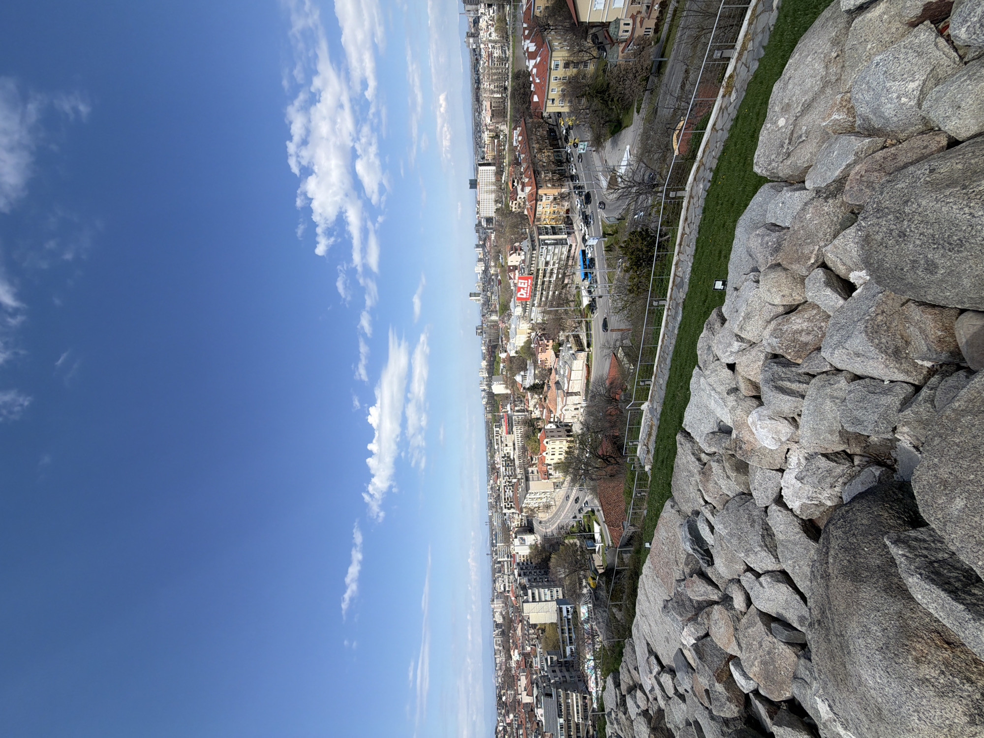 The image shows a view from Nebet Tepe, Plovdiv, Bulgaria. The foreground contains a stone wall and a patch of grass bordered by a metal fence. Behind the fence is a cityscape with buildings of varying heights and architectural styles. A street with vehicular traffic runs through the city. The background is dominated by a clear blue sky with scattered white clouds.