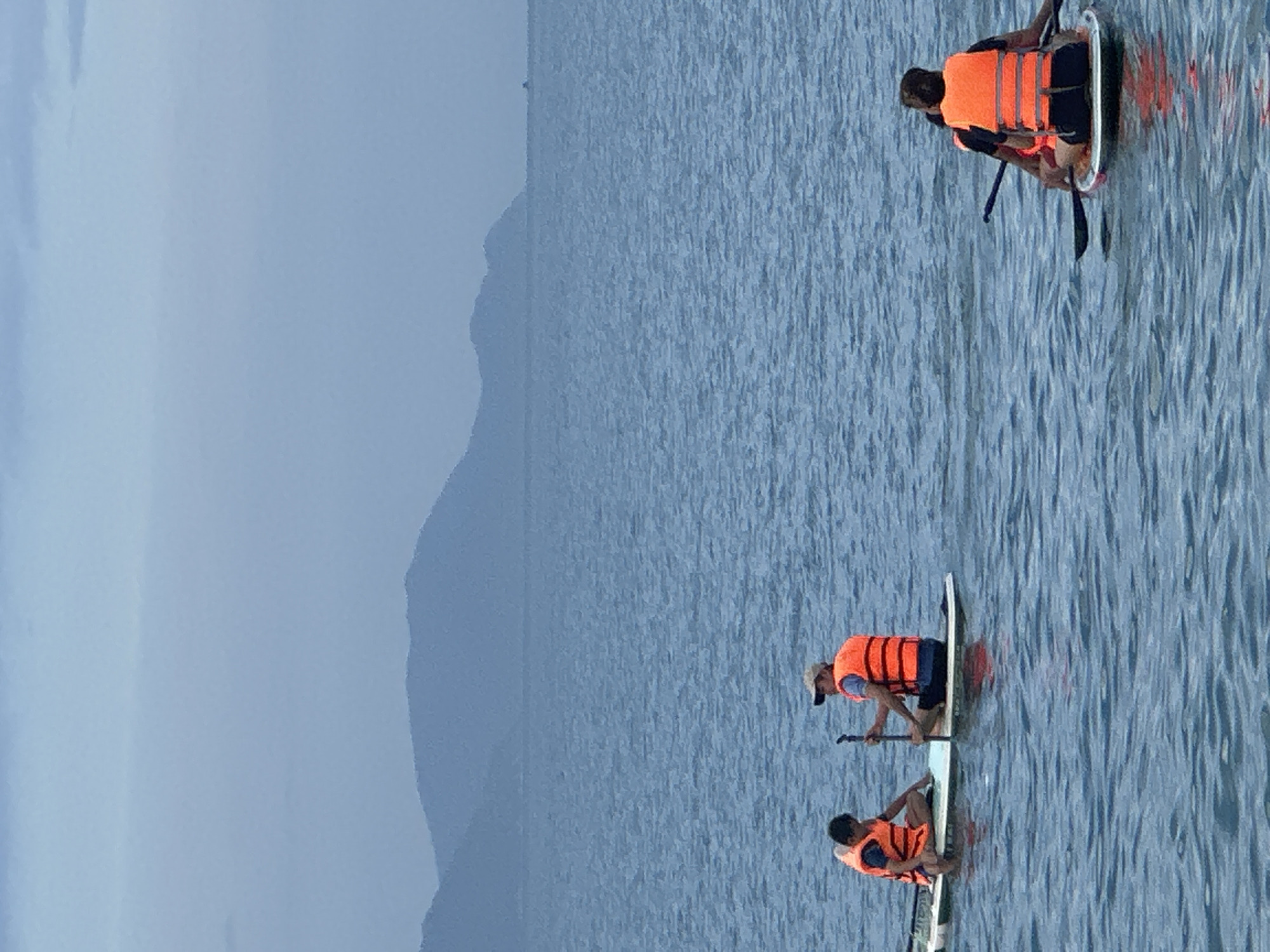 Three individuals are paddleboarding on a calm, blue-grey body of water under a muted sky. All three are wearing bright orange life vests, suggesting a focus on safety during the activity.
In the foreground, one person is seen sitting or kneeling on a paddleboard. Directly behind them, another person is either kneeling or standing on their board, holding a paddle and wearing a light-colored cap. Further in the background, a third person is also on a paddleboard, appearing to be kneeling or sitting with a paddle. The water's surface shows gentle ripples, indicating calm conditions.
The scene is set against a backdrop of distant, hazy blue-grey mountains that stretch across the horizon. The sky is a uniform, light blue-grey, blending subtly with the distant landscape and suggesting an overcast or slightly hazy day. The diffused lighting indicates it is daytime, possibly early morning or late afternoon, contributing to the serene and tranquil atmosphere of the scene. No discernible text is visible in the image. The context of Nha Trang Ward, Vietnam, suggests a coastal or bay environment, which aligns with the presence of a large body of water and mountains.