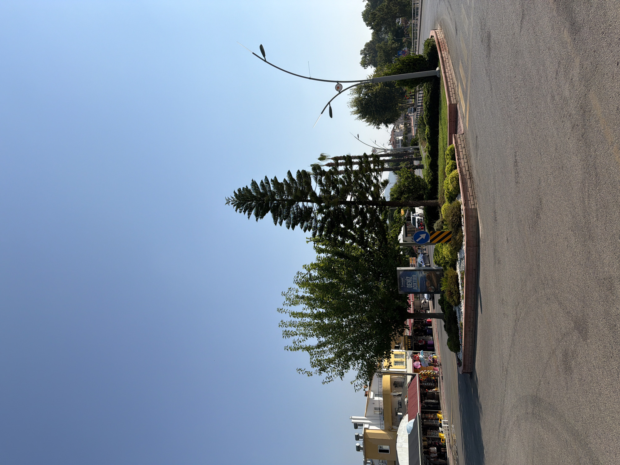 A sunny, clear day is depicted in this exterior shot of a traffic circle in Kemer, Turkey. The traffic circle is adorned with a cluster of trees, including a tall, conical evergreen and a leafy deciduous tree. Surrounding the trees is a neatly manicured garden bed with low-lying green shrubs and white decorative stones. A brick border defines the edge of the garden. A sign with Turkish text is visible, and a traffic sign indicating a right turn is also present. In the background, there are shops with awnings and buildings, some with solar panels on the roofs, suggesting a commercial and residential area. Streetlights with a distinctive curved design are also scattered throughout the scene. The road surface is asphalt, and there are yellow road markings. The sky is a vast expanse of clear blue.