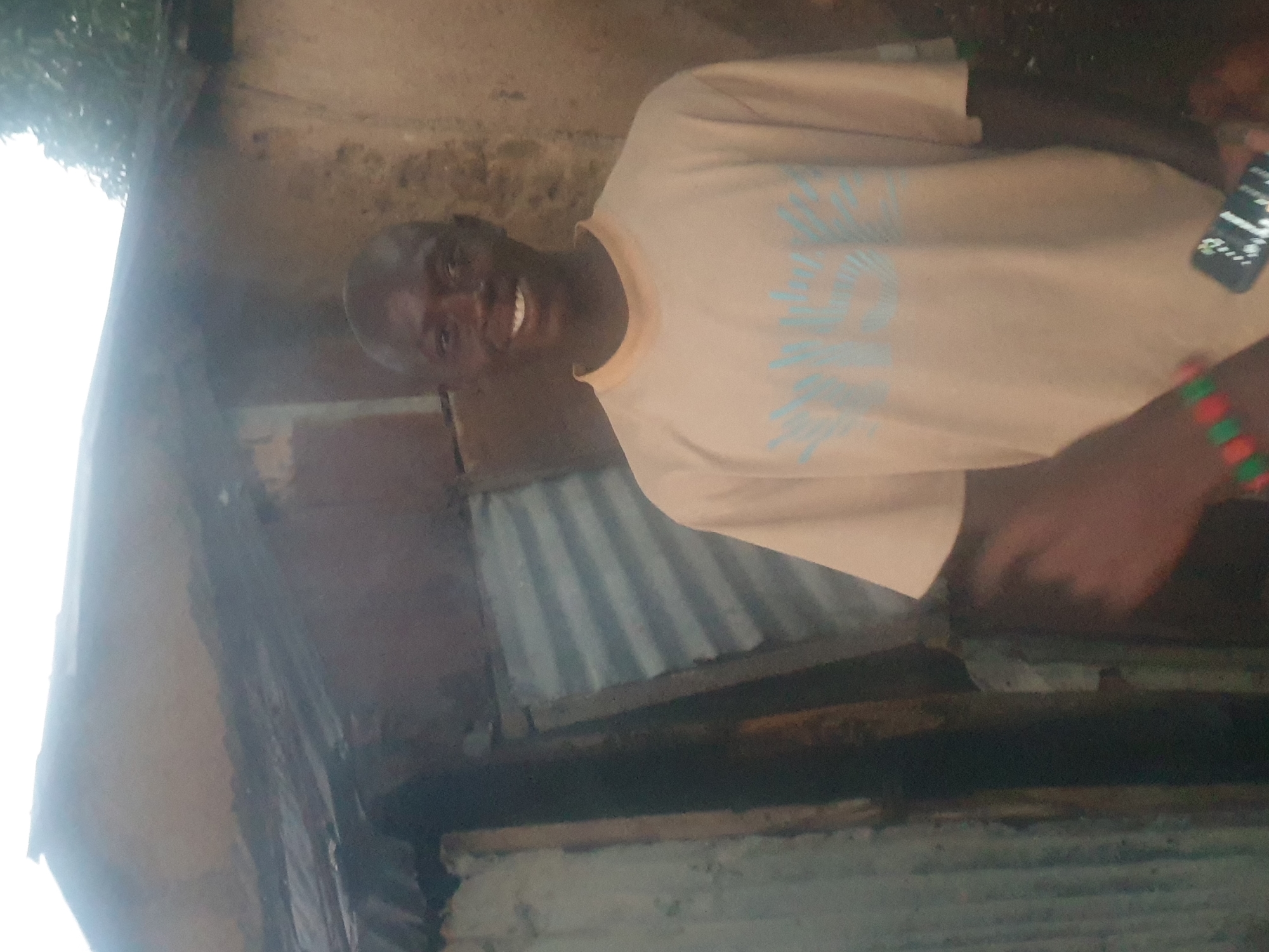 A man with dark skin, a shaved head, and a wide smile stands in front of a rustic building. He is wearing a light brown t-shirt with a blue graphic on the front. His left arm is visible, wearing a bracelet made of red and green beads. In his left hand, he holds a dark-colored mobile phone with a white screen displaying a few icons. The building behind him appears to be constructed with corrugated metal and possibly some wooden elements. The overall environment suggests an outdoor setting. The image does not provide any information about the location being in the city of Washington, United States of America.