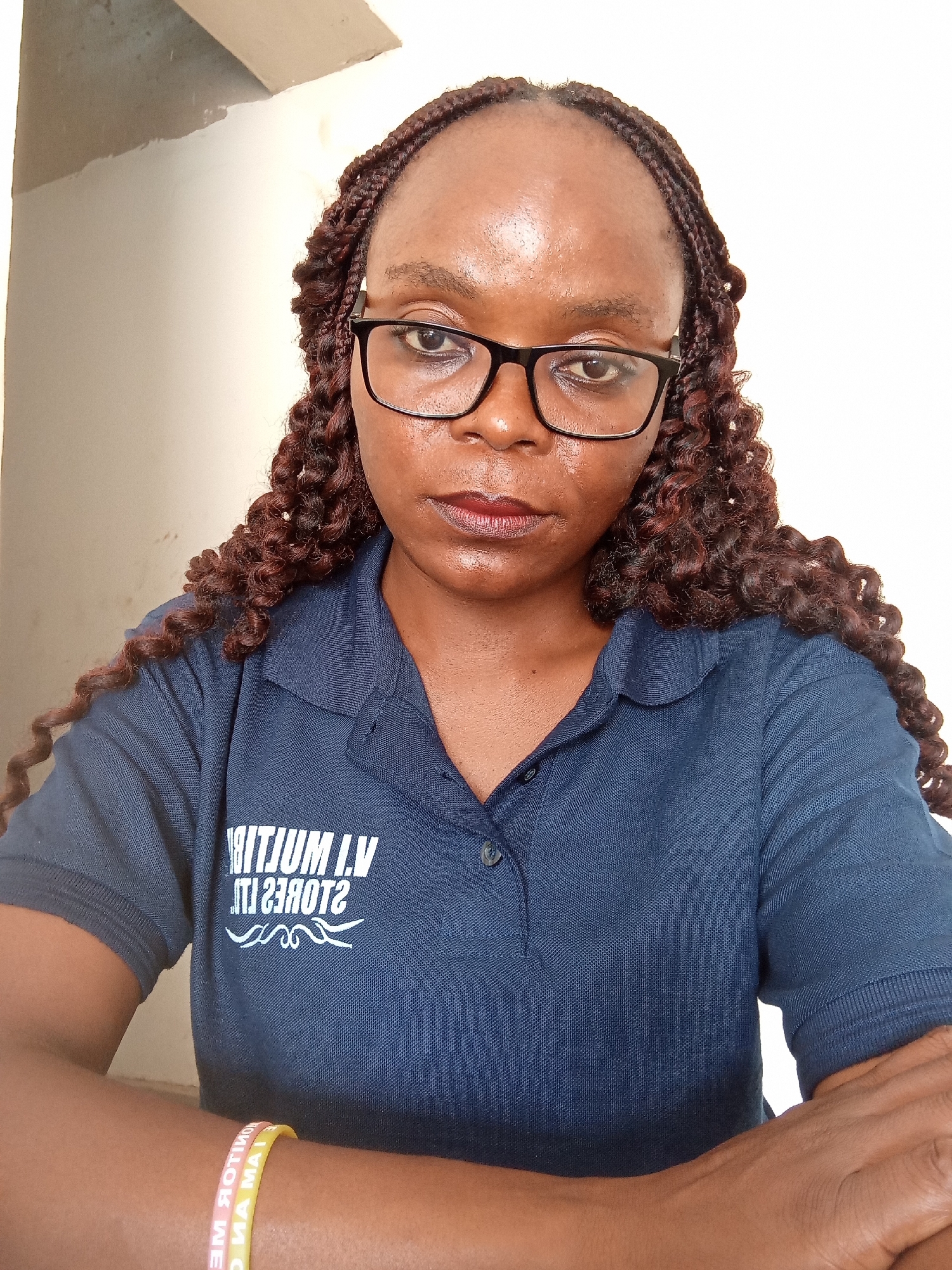 The media file shows a close-up, waist-up, indoor portrait of a Black woman wearing glasses and a navy blue collared shirt. Her hair is styled in dark brown braids. She is looking directly at the camera with a neutral expression. The background is a plain, light-colored wall, with a textured, beige ceiling visible in the upper left corner.
On her shirt, there is a white logo that appears to read "V.I. MULTIJUM L.V." with additional lettering underneath that is difficult to discern. The woman is wearing two wristbands, one yellow and one pink, on her left forearm. The pink wristband has white text that is partially visible and appears to read "ME WOK" and "MA MAI". The yellow wristband also has white text, but it is less legible.
The lighting suggests an indoor setting, possibly during daylight hours, as there is no indication of artificial lighting. The overall impression is a casual, self-taken portrait.